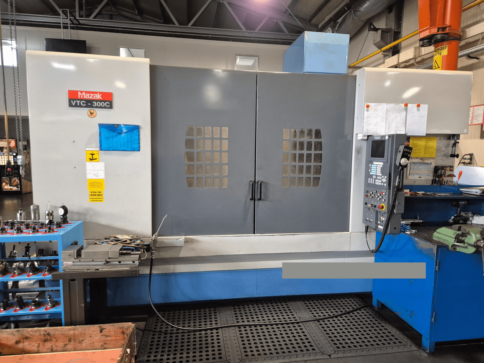 Mazak VTC-300C Maschine in einer Fabrik, Vorderansicht mit Bedienfeld, grauen Türen mit Fenstergitter und nahe gelegenen Geräten.