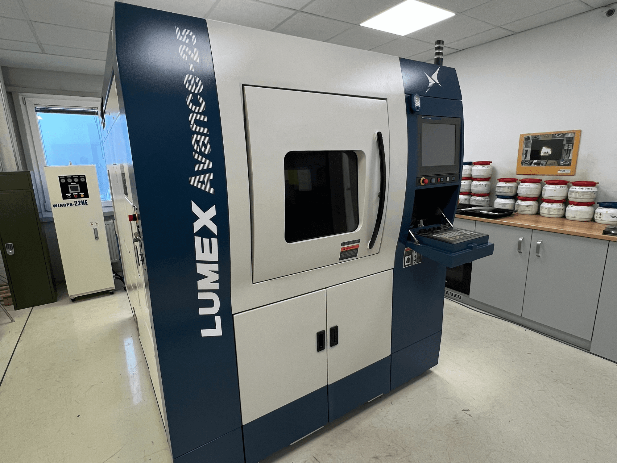 LUMEX Avance-25 3D-Drucker in einer Laborumgebung, Vorderansicht. Bedienfeld mit Bildschirm und Tasten sichtbar, Geräte im Hintergrund.