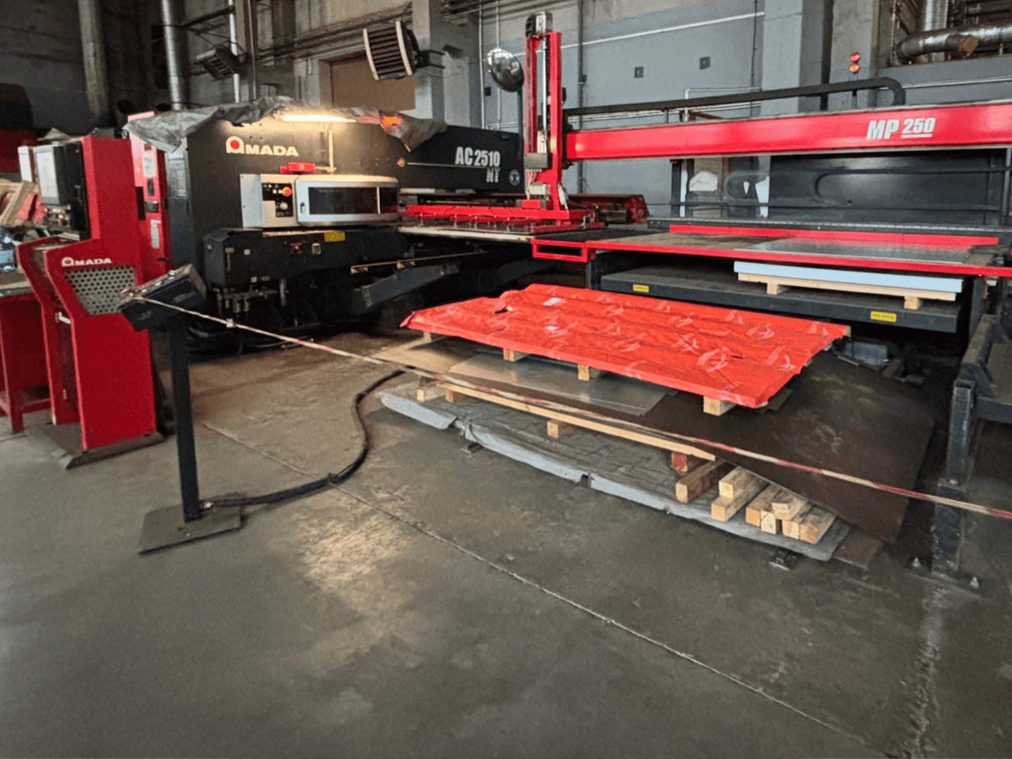Amada AC2510 NT Laserschneider in einer Fabrikhalle, Vorderansicht, schwere Maschinen mit Blechen und Paletten auf dem Boden.