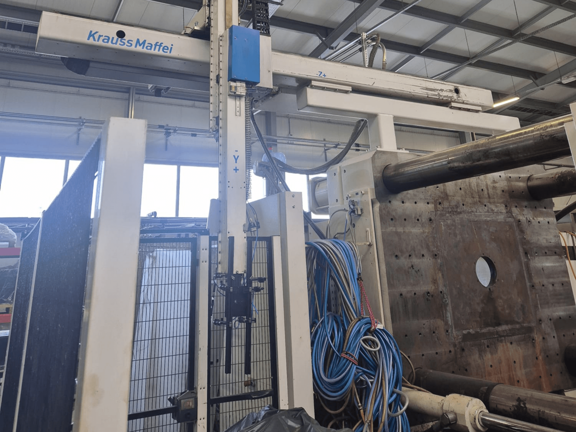 Hydraulische Spritzgießmaschine Krauss Maffei 1300-12000 MX, Vorderansicht mit mechanischen Armen und Hydraulikanschlüssen.