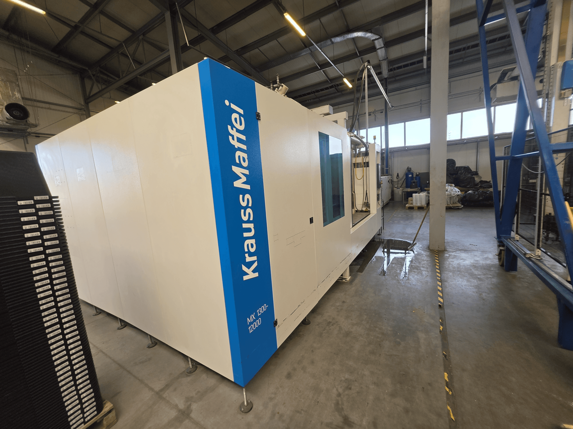 Hydraulische Spritzgießmaschine vom Typ Krauss Maffei MX 1300-12000 in der Seitenansicht, die ihr großes weißes Fahrgestell und das blaue Markenlogo zeigt.