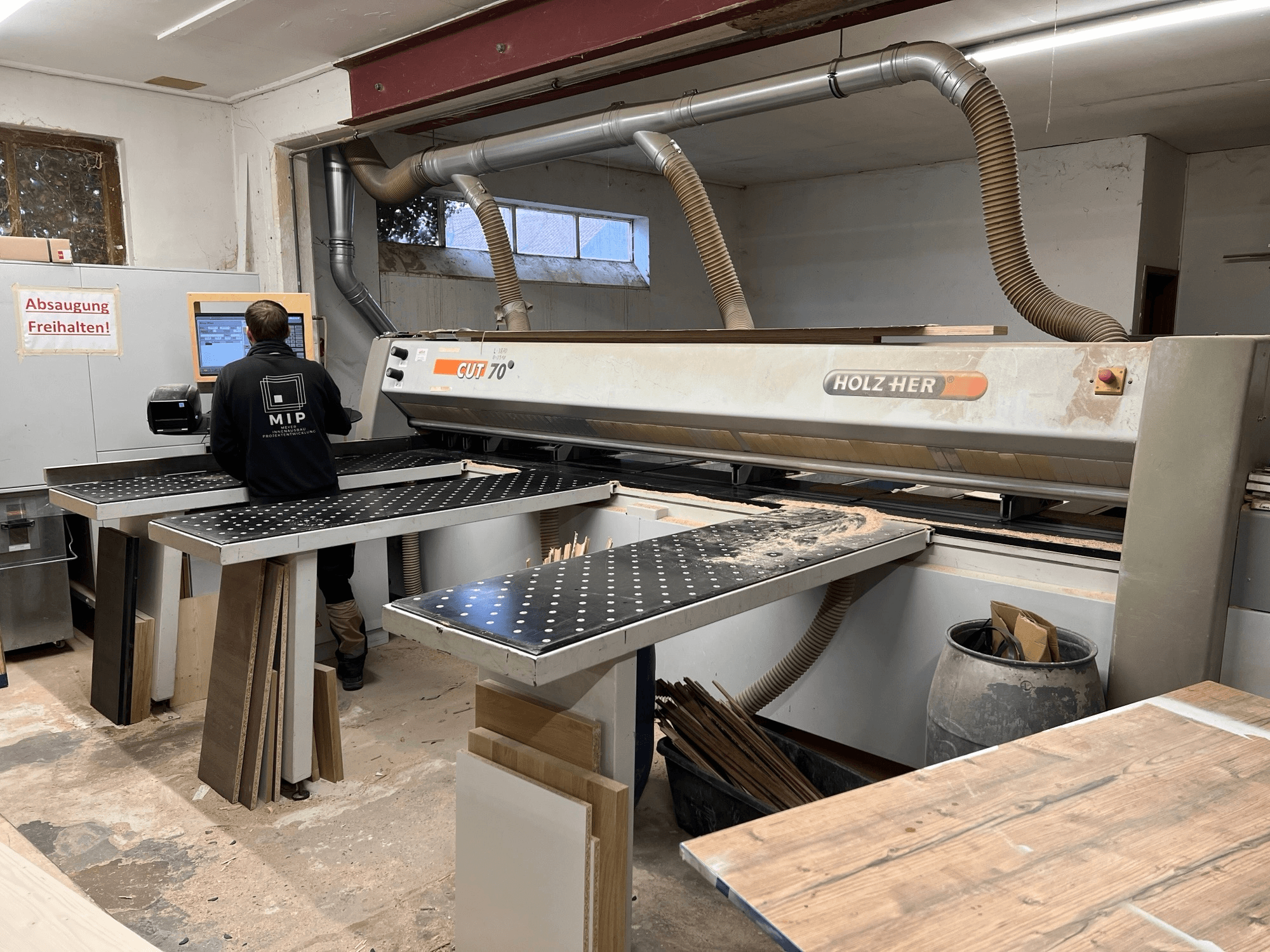 Holzher CUT 70 Säge in einer Werkstatt, von der Seite gezeigt, mit einem Arbeiter, der die Steuerungen bedient und Holzplatten auf den Tischen.