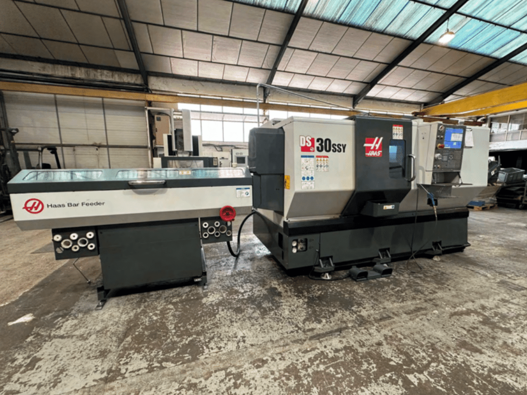 Haas DS-30SSY CNC-Drehmaschine mit Haas Stangenlader, Seitenansicht mit Bedienfeld und Vorschubmechanismus in einer Werkstattumgebung.