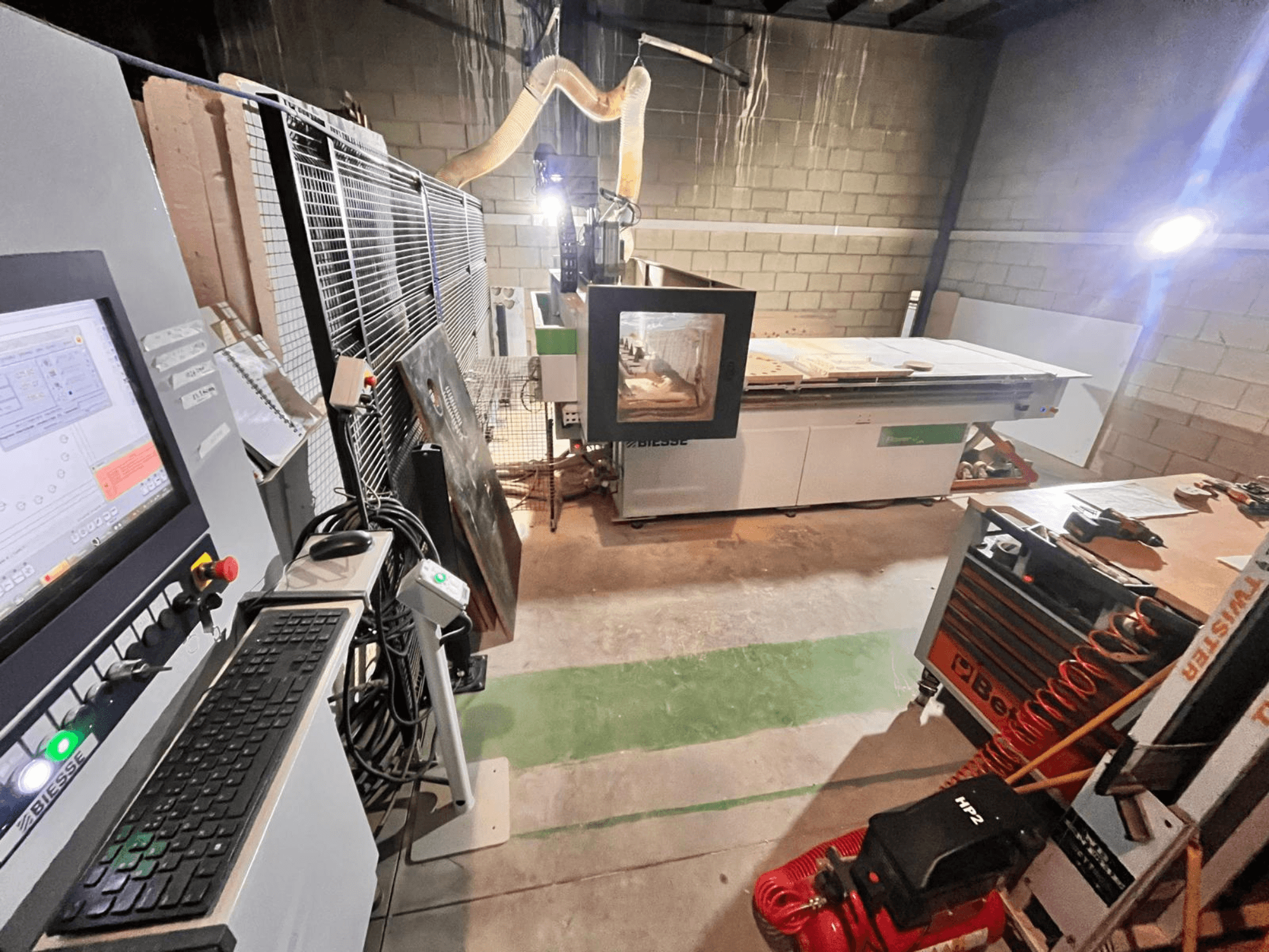 Biesse CNC-Maschine in einer Werkstatt, Seitenansicht mit Bedienfeld, Staubabsaugung, Werkzeugen und verschiedenen Materialien auf dem Tisch.