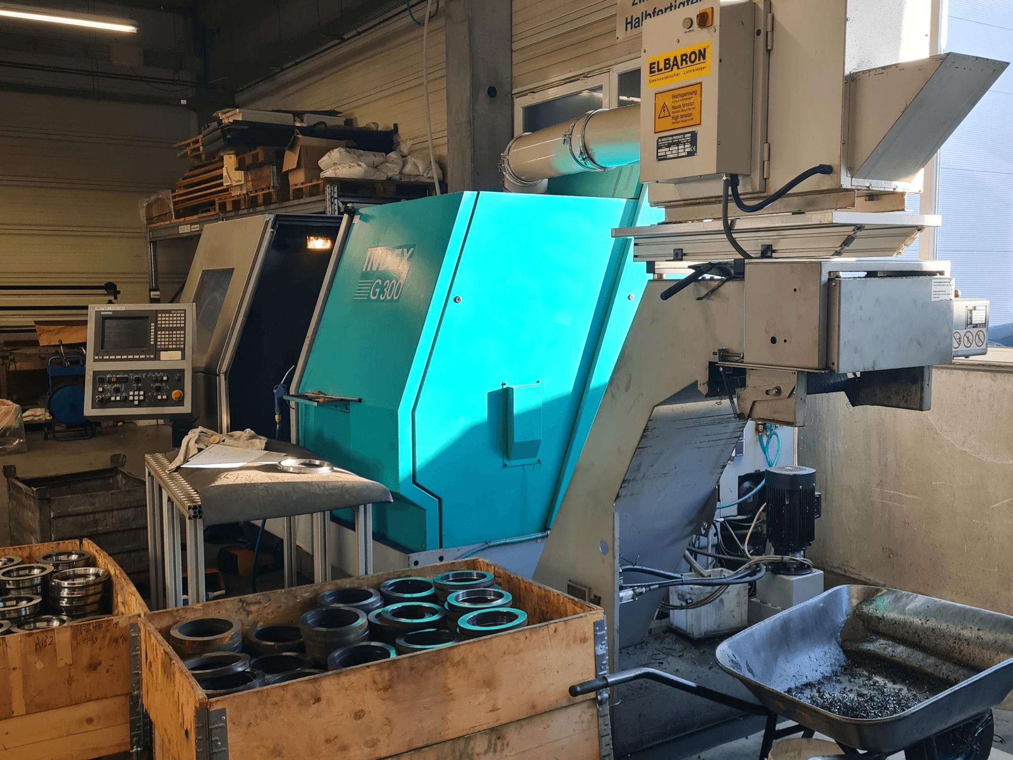 Vorderansicht der Horizontal-Drehmaschine „Index G300 RatioLine“ mit Bedienfeld, umgeben von Werkstücken und einem Förderband.