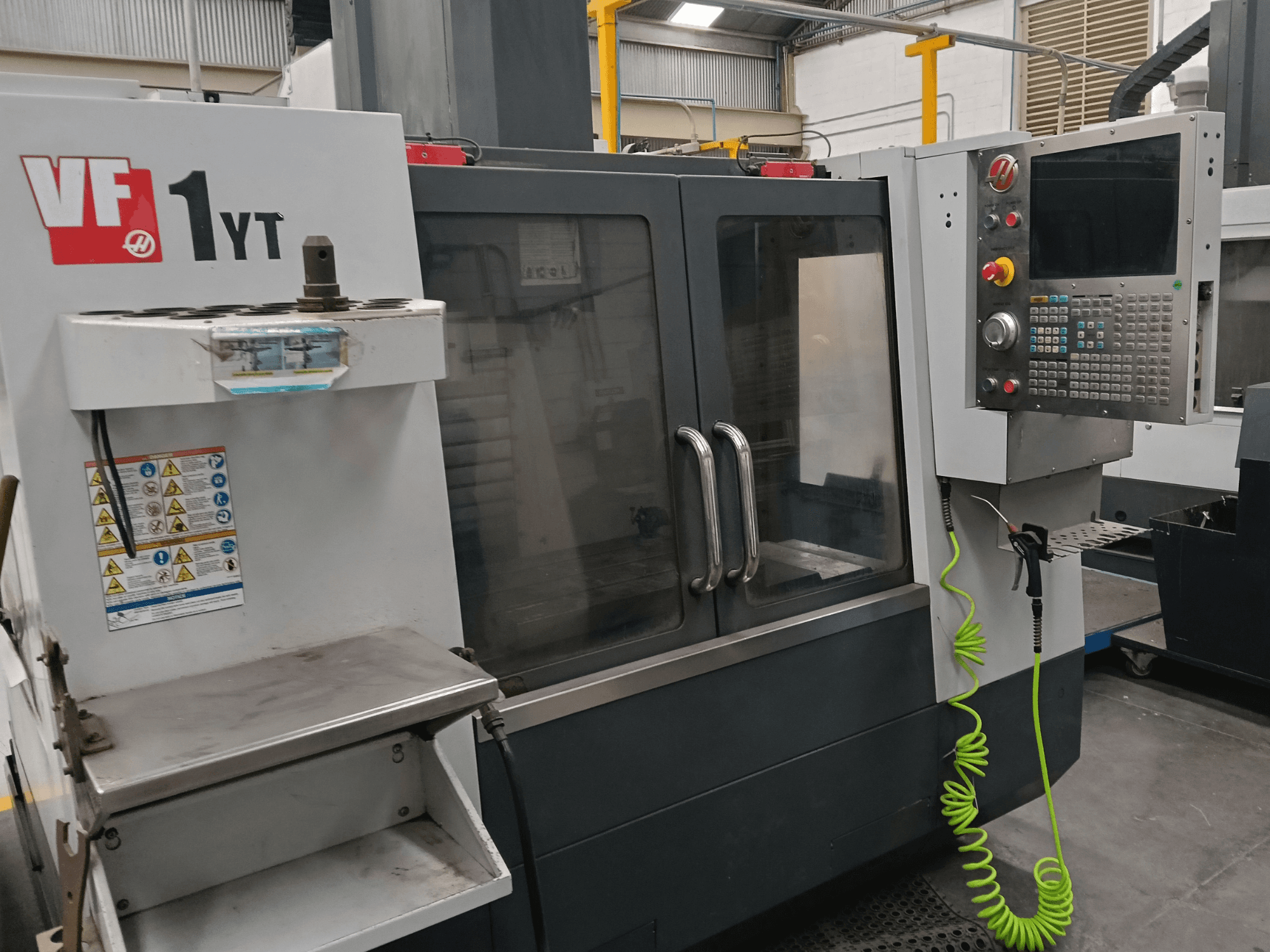 Vertikales Bearbeitungszentrum HAAS VF-1YT-2, Vorderansicht, mit Bedienfeld, Werkzeughalter, Sicherheitsaufklebern und Bearbeitungsbereich.
