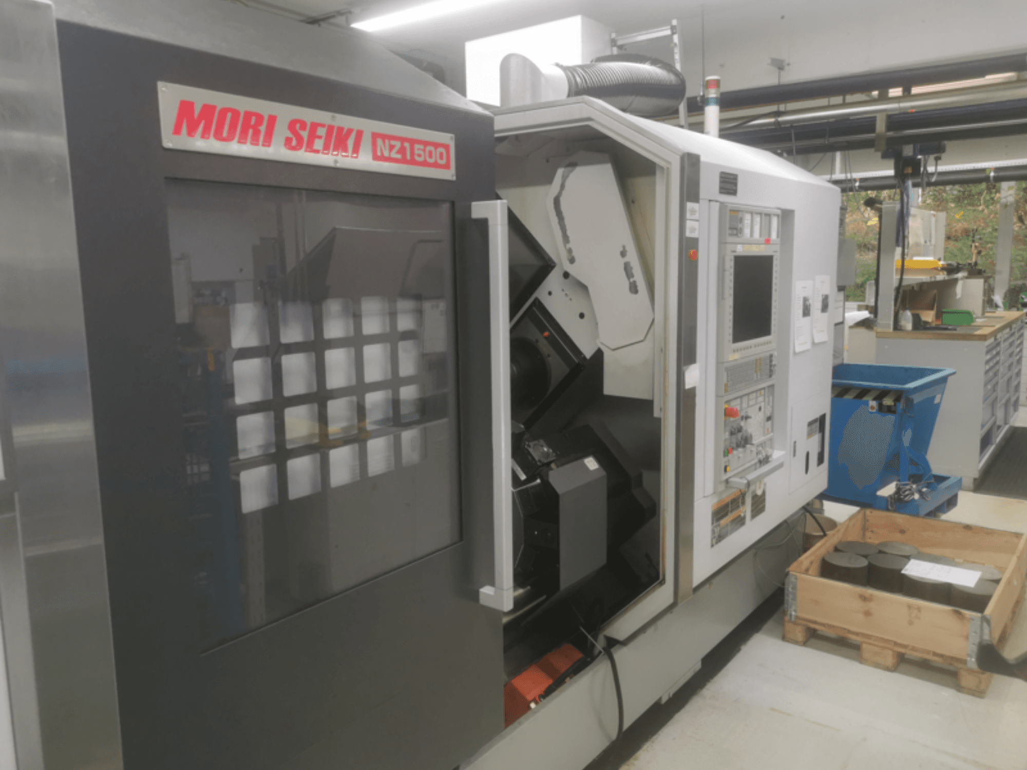 MORI SEIKI NZ1500 CNC-Maschine in einem leichten Winkel, zeigt Bedienfeld und Arbeitsbereich mit Metallwerkstücken auf einer Holzpalette.