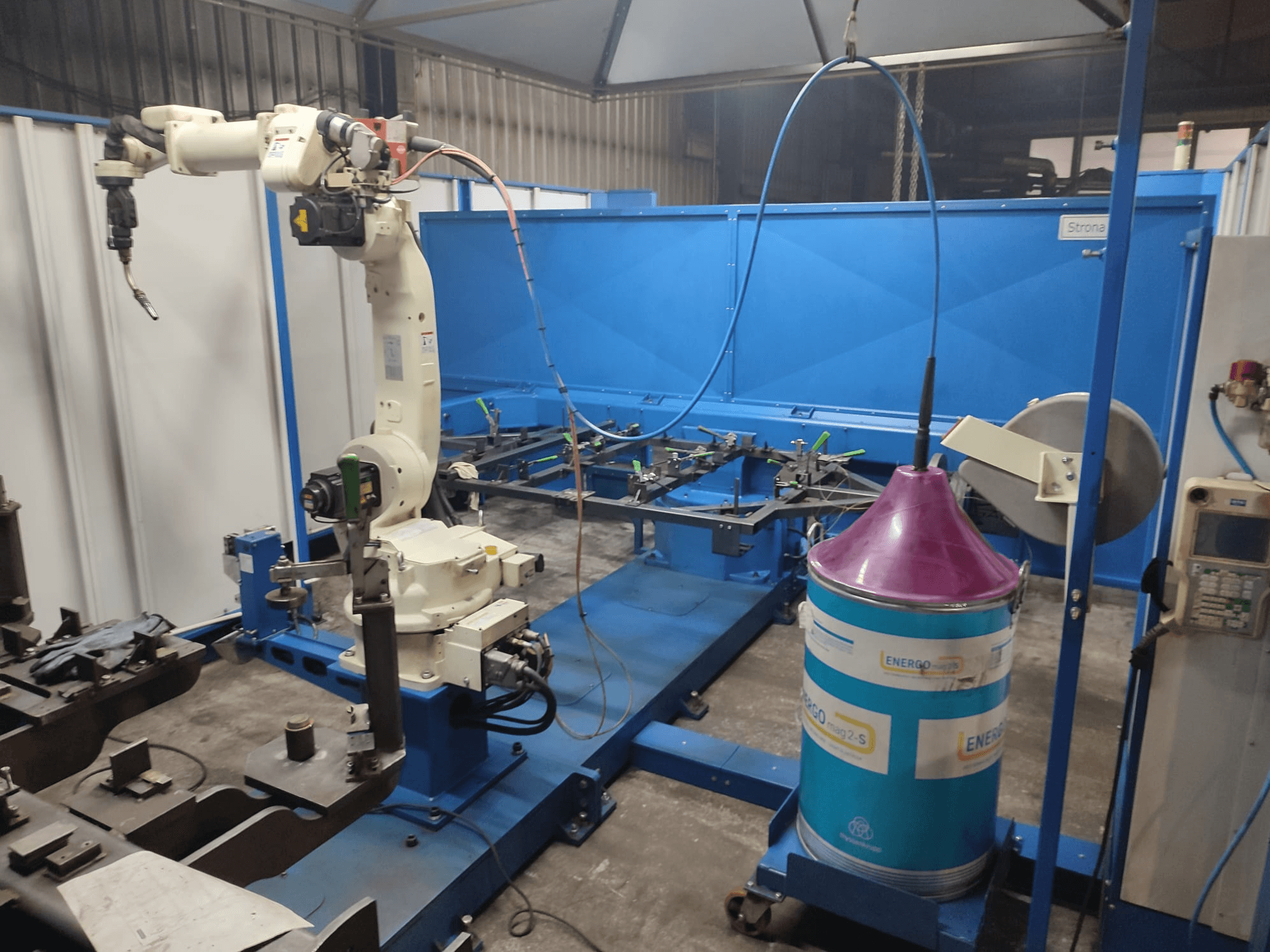 Industrieller Roboterarm mit weißem und blauem Sockel, eingerichtet in einer Werkstatt mit blauer Arbeitsfläche und Metallteilen.