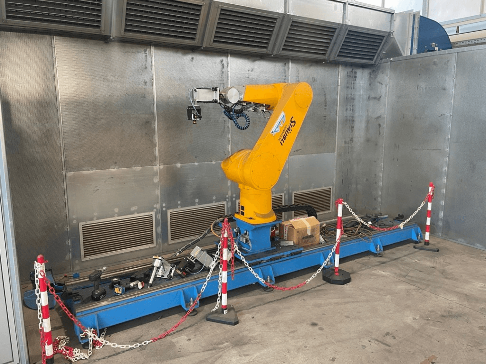 STAUBLI CS8C-RX160 Roboterarm in einer Werkstatt, Seitenansicht, mit Kabeln und verschiedenen Werkzeugen auf einem blauen Sockel, umgeben von Schutzwänden.