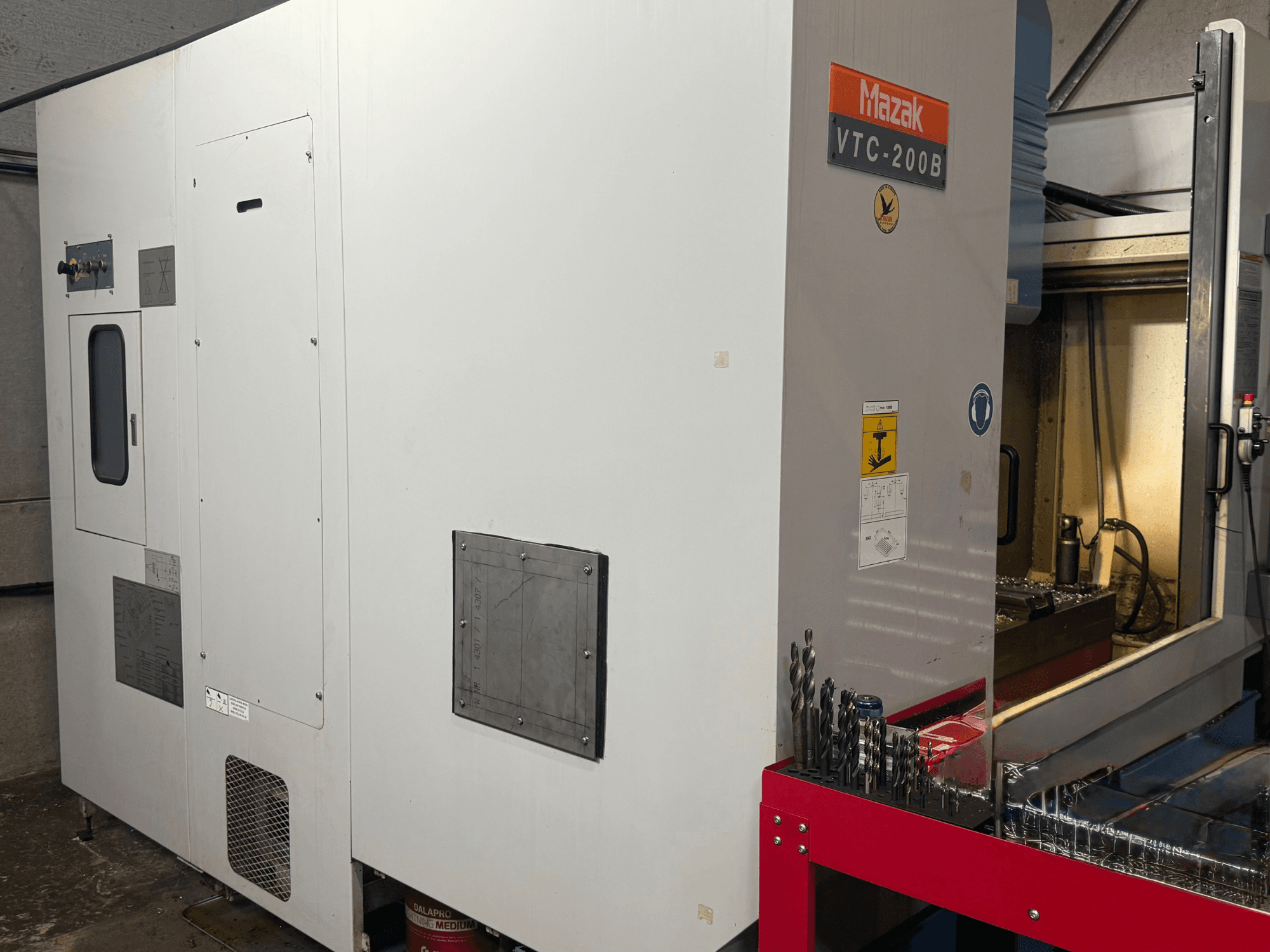 Vertikales Bearbeitungszentrum Mazak VTC-200B in Seitenansicht, mit beschrifteten Bedienelementen und Bohrern auf einem roten Aufbewahrungsregal.