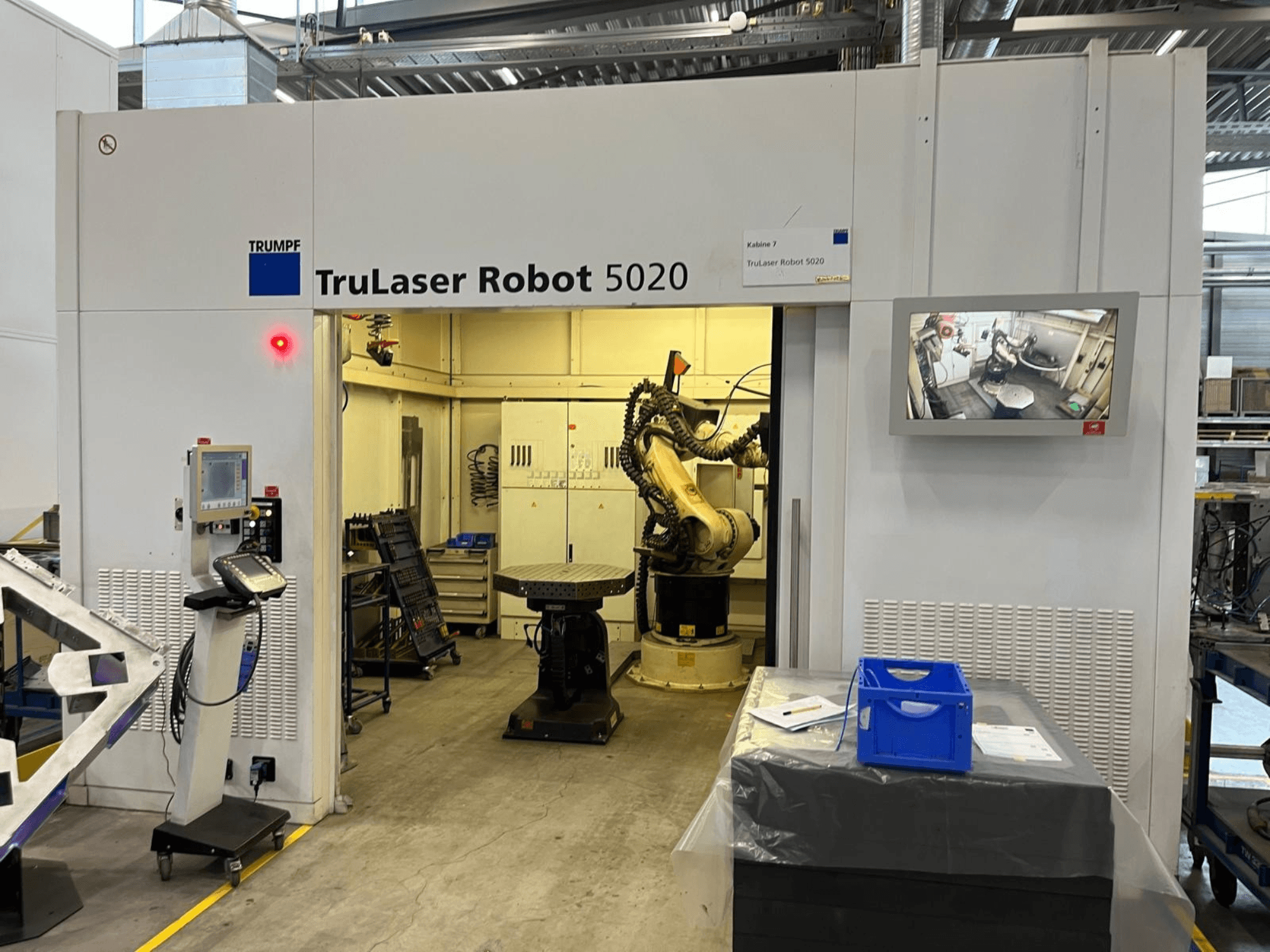 TRUMPF TruLaser Robot 5020 in einer industriellen Umgebung, mit Bedienelementen, einem Roboterarm und einem Monitor, auf dem die Abläufe angezeigt werden.