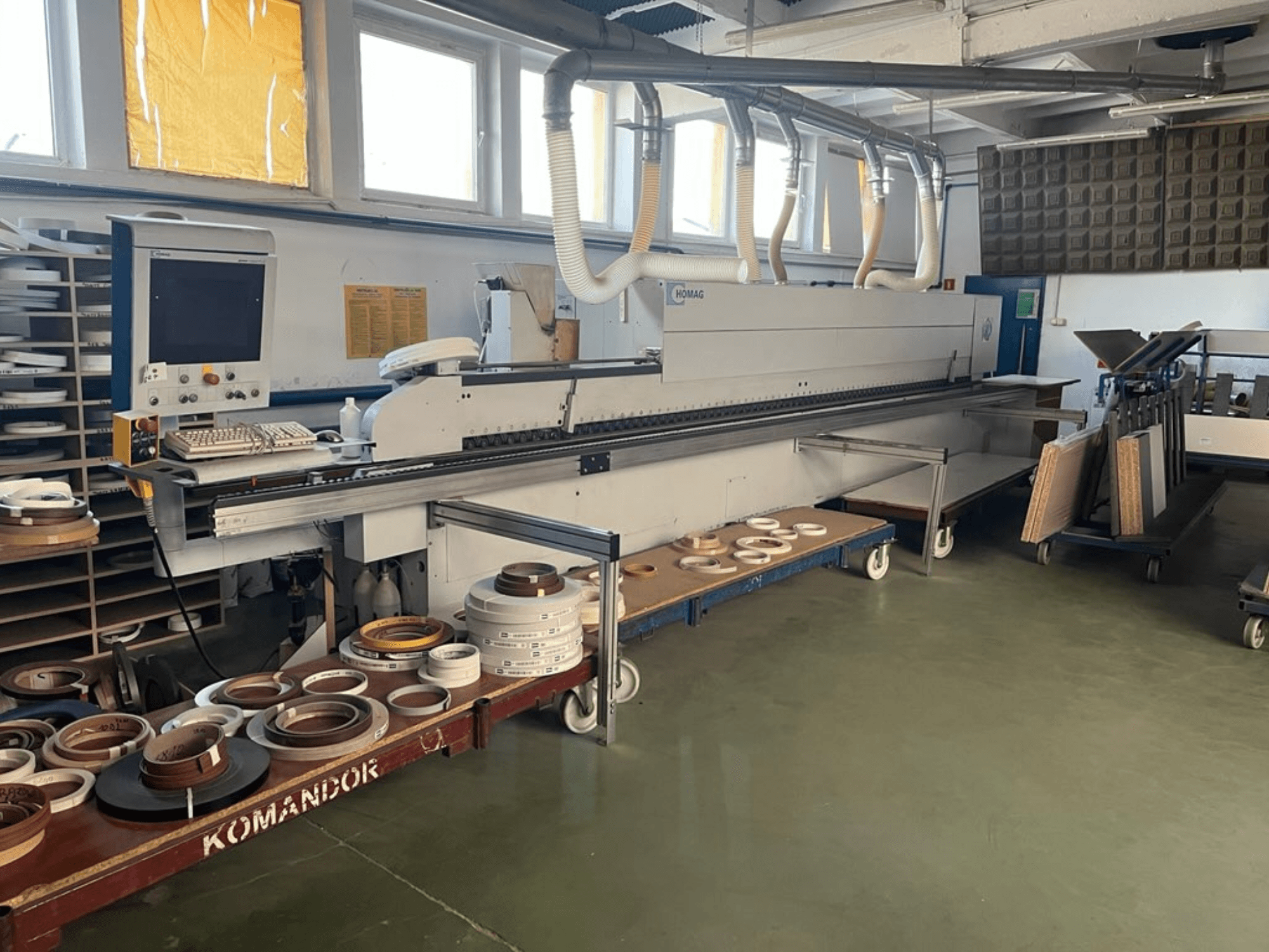 HOMAG Kantenanleimmaschine in einer Werkstatt, mit Bedienelementen und verschiedenen Kantenanleimmaterialien auf einem blauen Wagen. Seitenansicht.