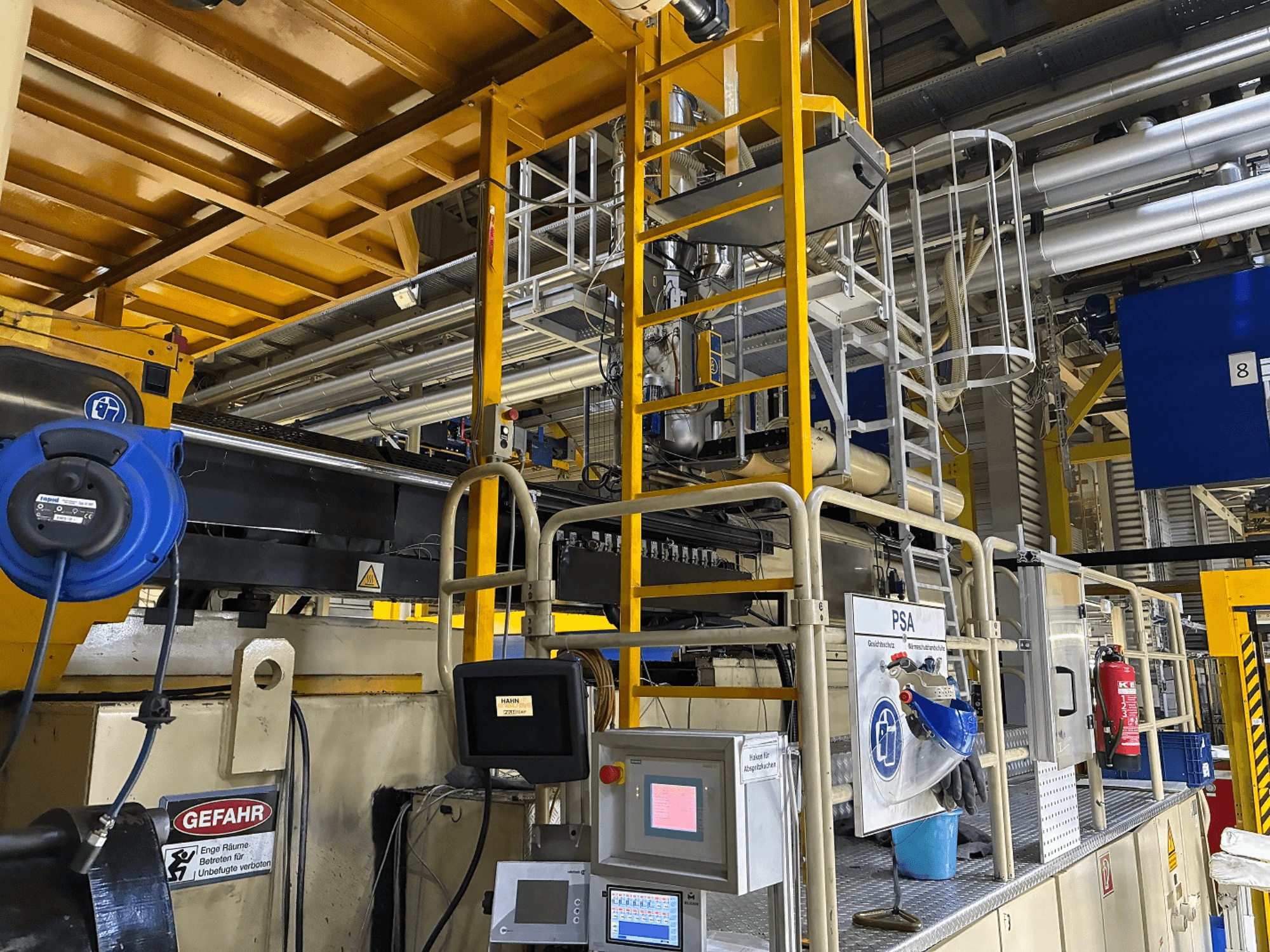 Schalttafel einer Industriemaschine mit blauer Sicherheitsausrüstung und gelbem Gerüst, Seitenansicht mit Rohren und Warnschildern.