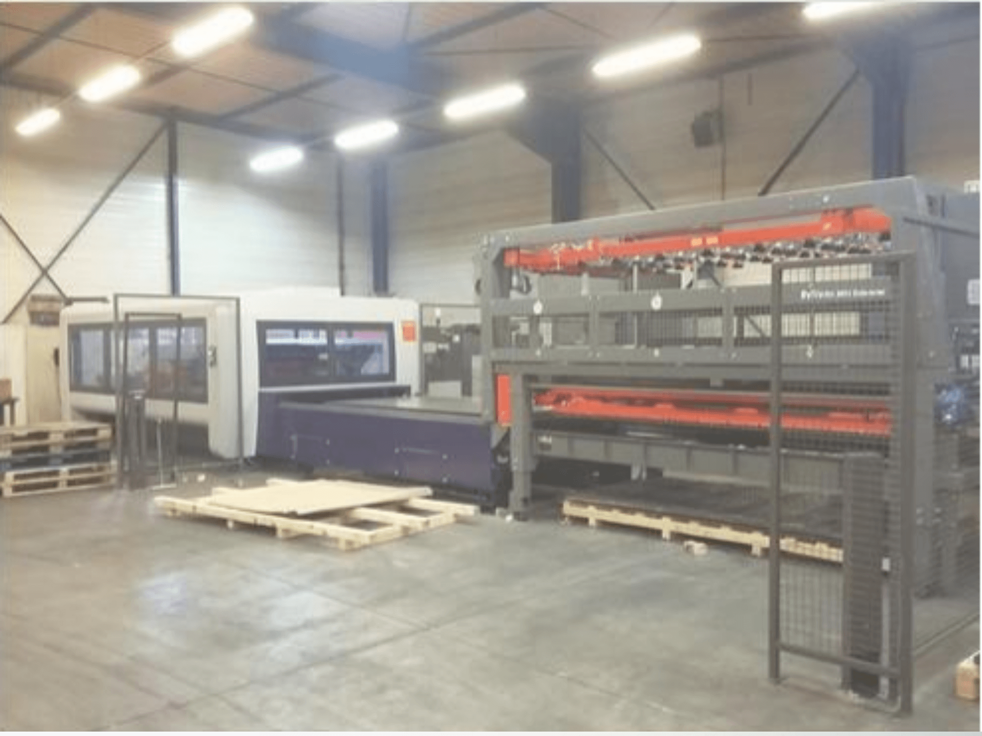 Aufstellung von Industriemaschinen in einem Lagerhaus, darunter ein grauer Laserschneider und eine violette CNC-Fräse, Vorderansicht mit Paletten.