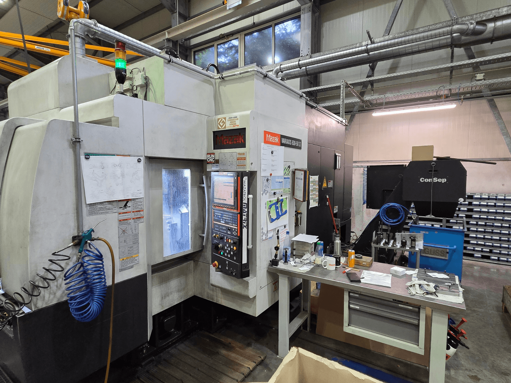 Mazak Variaxis 630-5X CNC-Maschine in einer Werkstatt, Vorderansicht mit Bedienfeld, Schlauch und Zubehör auf dem Seitentisch.