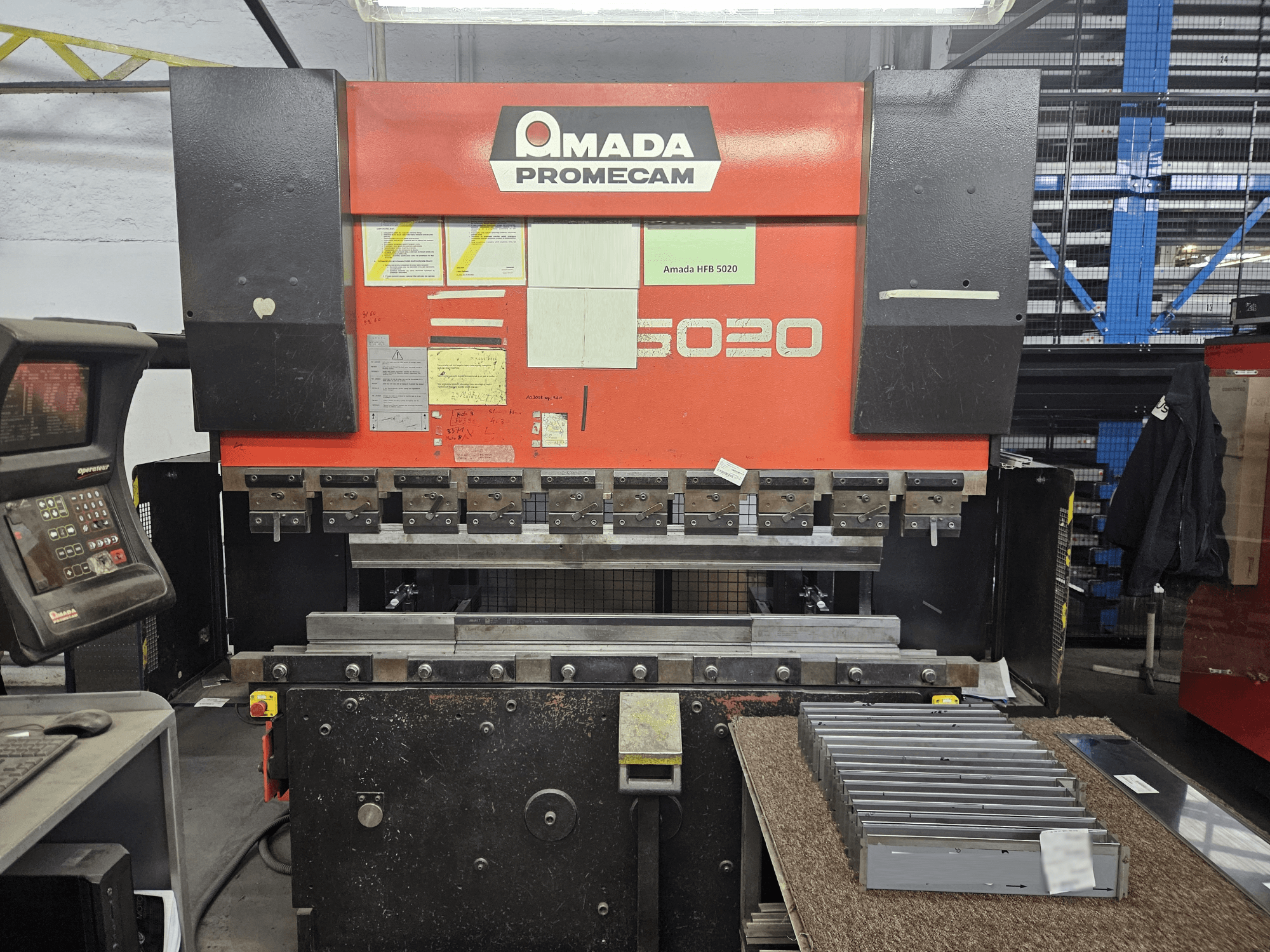 Amada HFBO 5020 Abkantpresse in Vorderansicht, mit Bedienfeld und gestapelten Metallteilen auf der Arbeitsfläche.