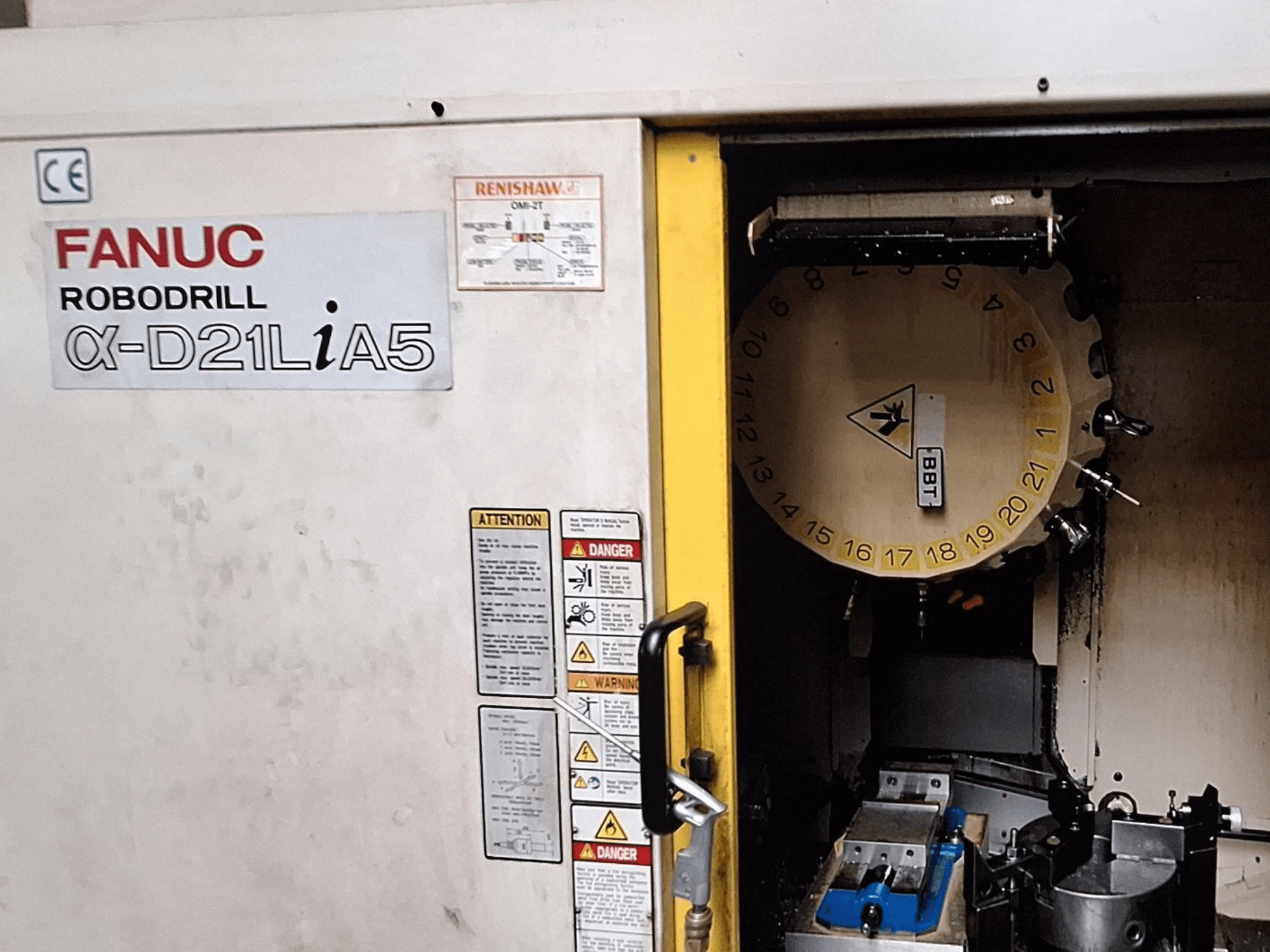 FANUC Robodrill α-D21LiA5, Vorderansicht. Zeigt das Werkzeugkarussell und den Bearbeitungsbereich mit Klemmen und Warnhinweisen an der Tür.