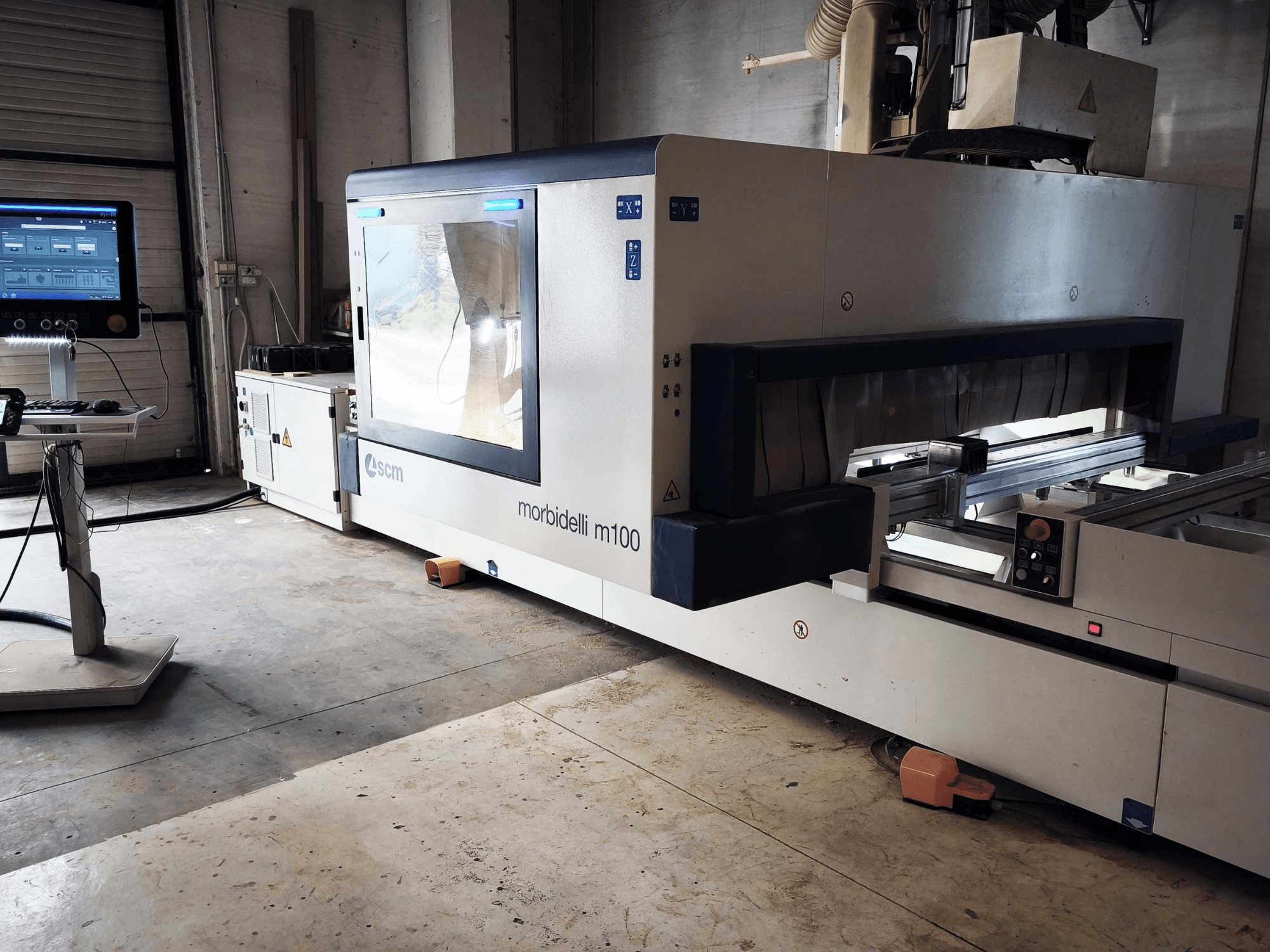 CNC-Holzbearbeitungszentrum SCM Morbidelli M 100 in Seitenansicht, mit Blick auf das Bedienfeld, den Arbeitsbereich und die Sicherheitsvorrichtungen.
