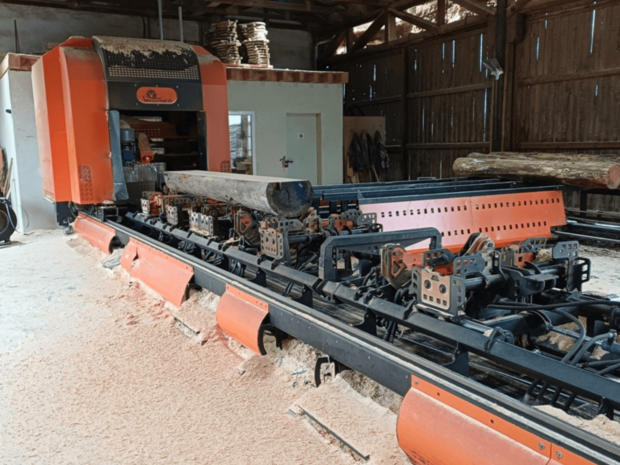 Orangefarbene Holzfräsmaschine mit einem auf dem Förderband positionierten Holzstamm, von vorne gesehen in einer Werkstattumgebung.