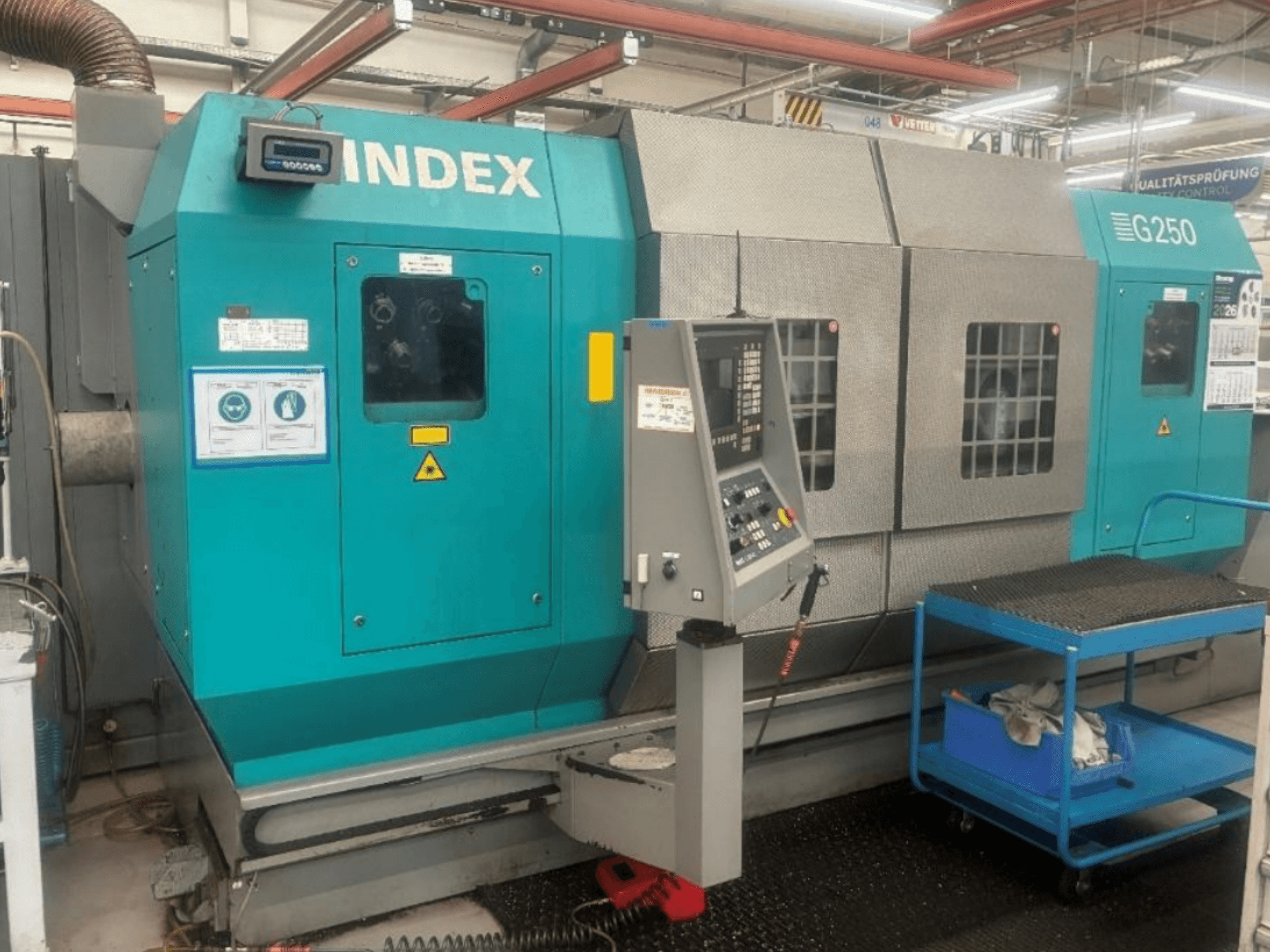 Horizontal-Drehmaschine INDEX G250, Vorderansicht mit Bedienfeld und Metallgehäuse, mit Sicherheitsaufklebern und einem blauen Transportwagen.