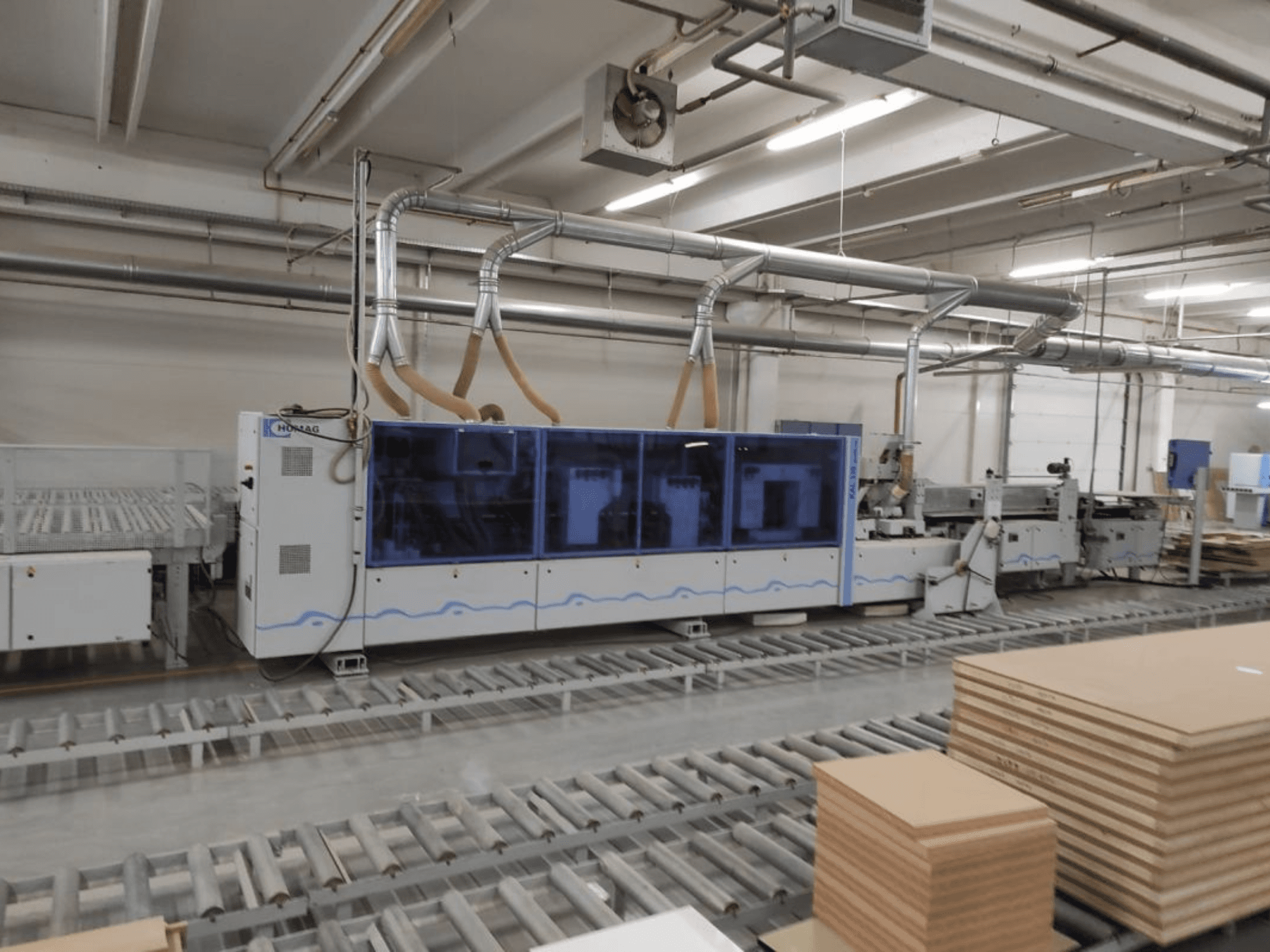 HOMAG Produktionsmaschine in einer Fabrik, Vorderansicht, mit blauem Gehäuse und Förderanlage mit gestapelten Sperrholzplatten.