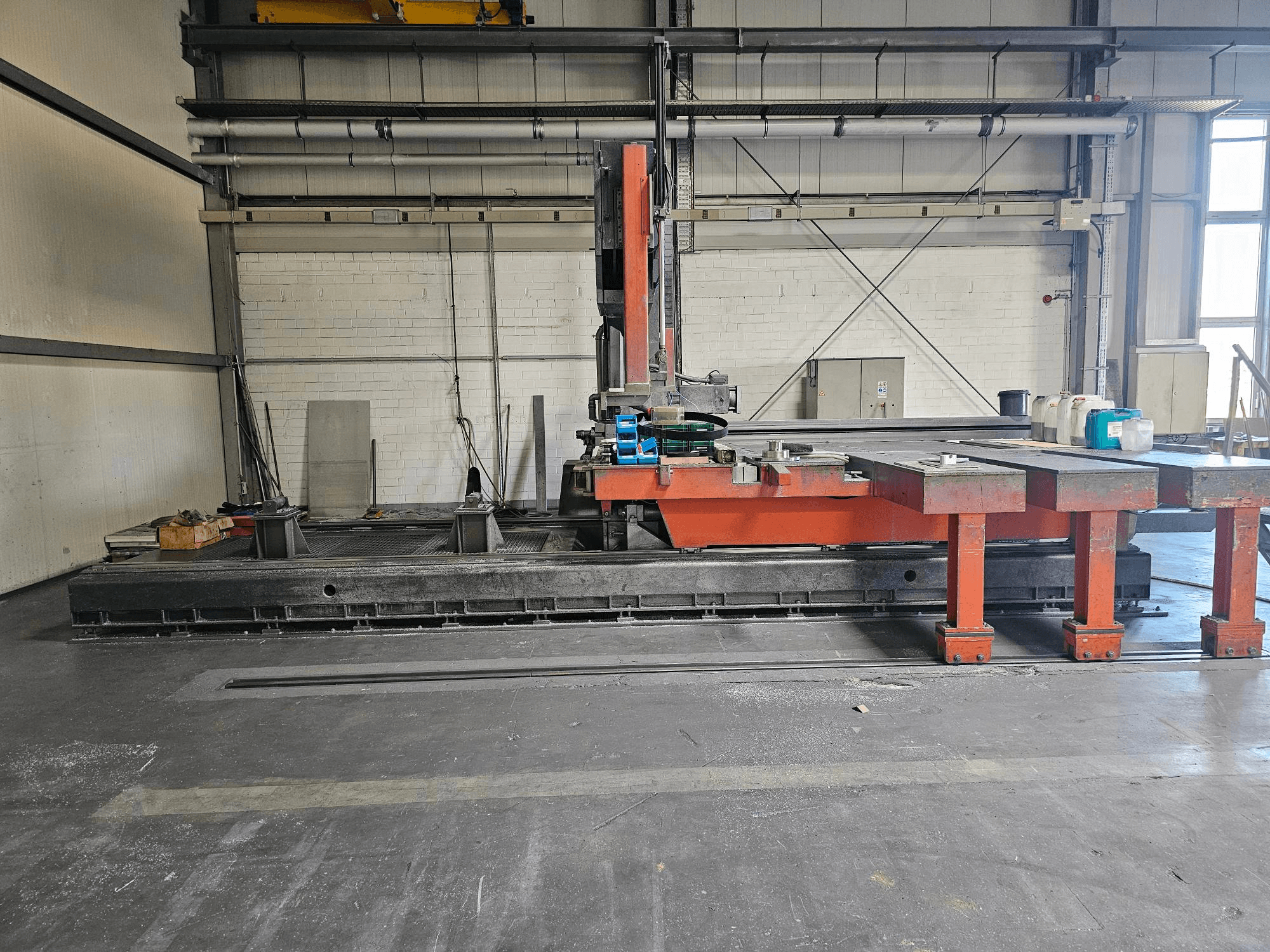 Industrielle Schneidemaschine in einer offenen Werkstatt, orange und schwarz, mit einem großen Tisch; Seitenansicht mit Zubehör und Arbeitsbereich.