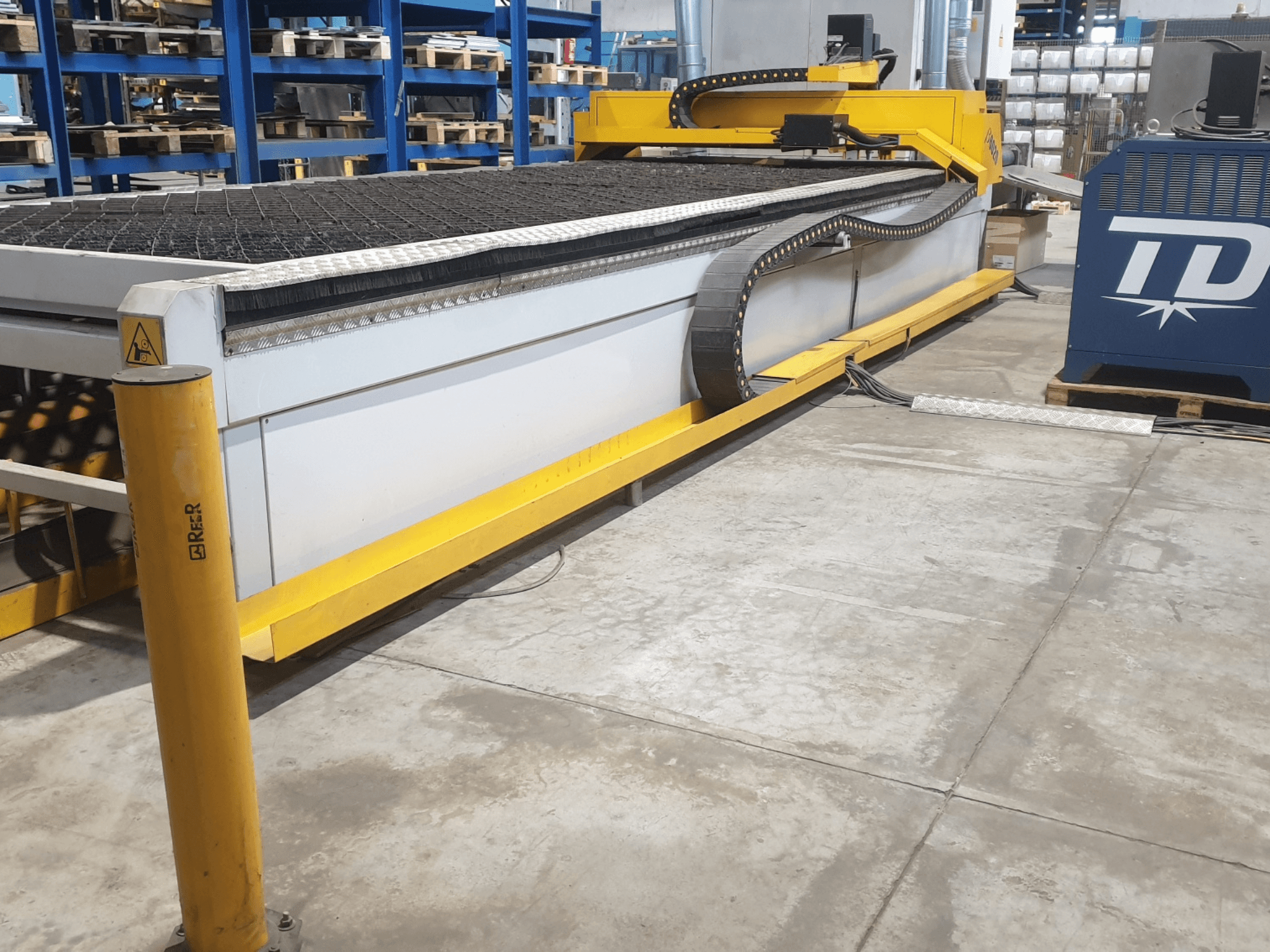 Industrielle Schneidemaschine mit gelber Verkleidung, Vorderansicht, mit strukturiertem Bett und Bedienfeld, aufgestellt in einer Fabrikhalle.