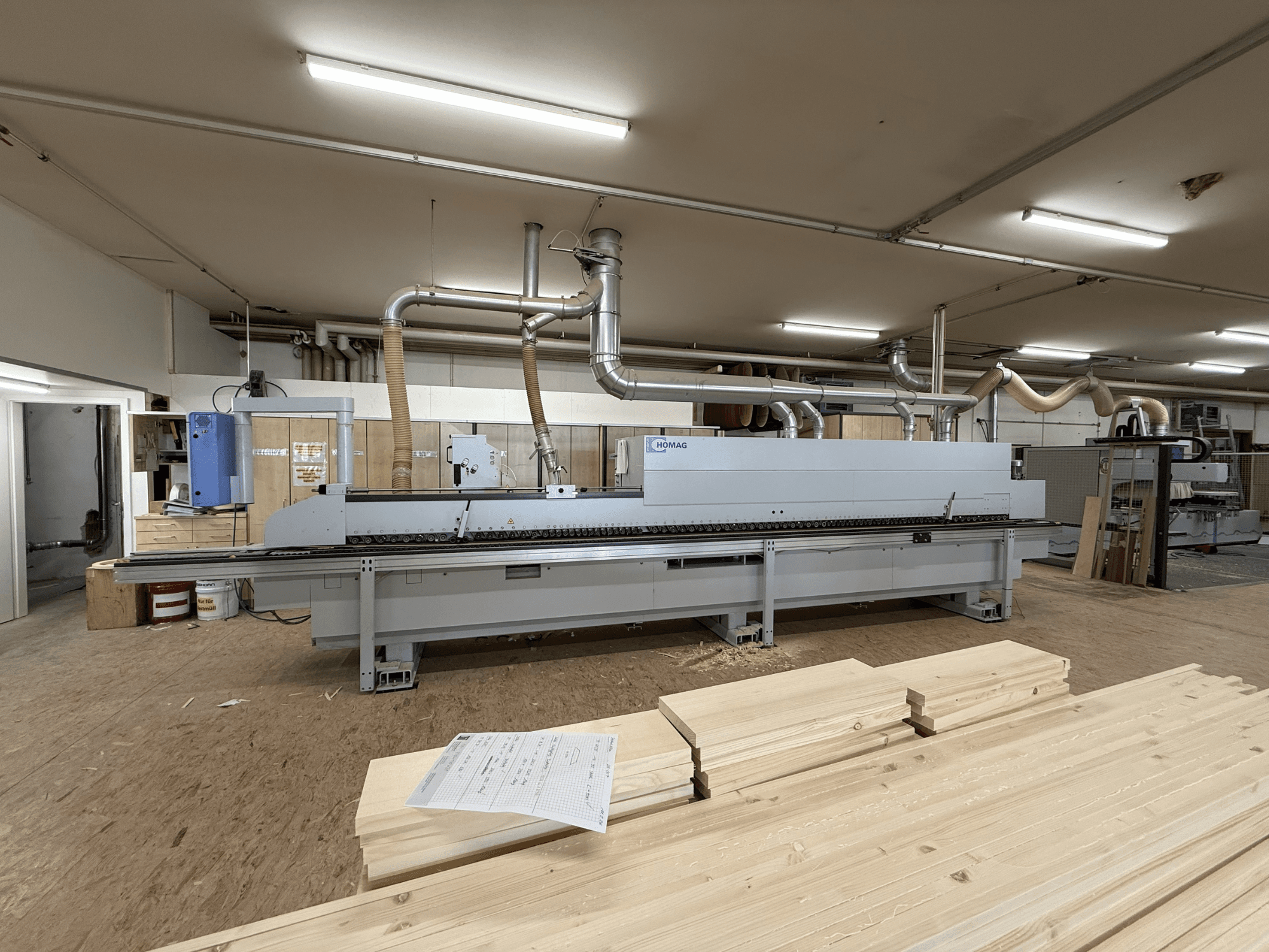 HOMAG KAL211 Kantenanleimmaschine in einer Werkstatt, Vorderansicht, mit Holzplatten und Maschinen in einer gut beleuchteten Umgebung.
