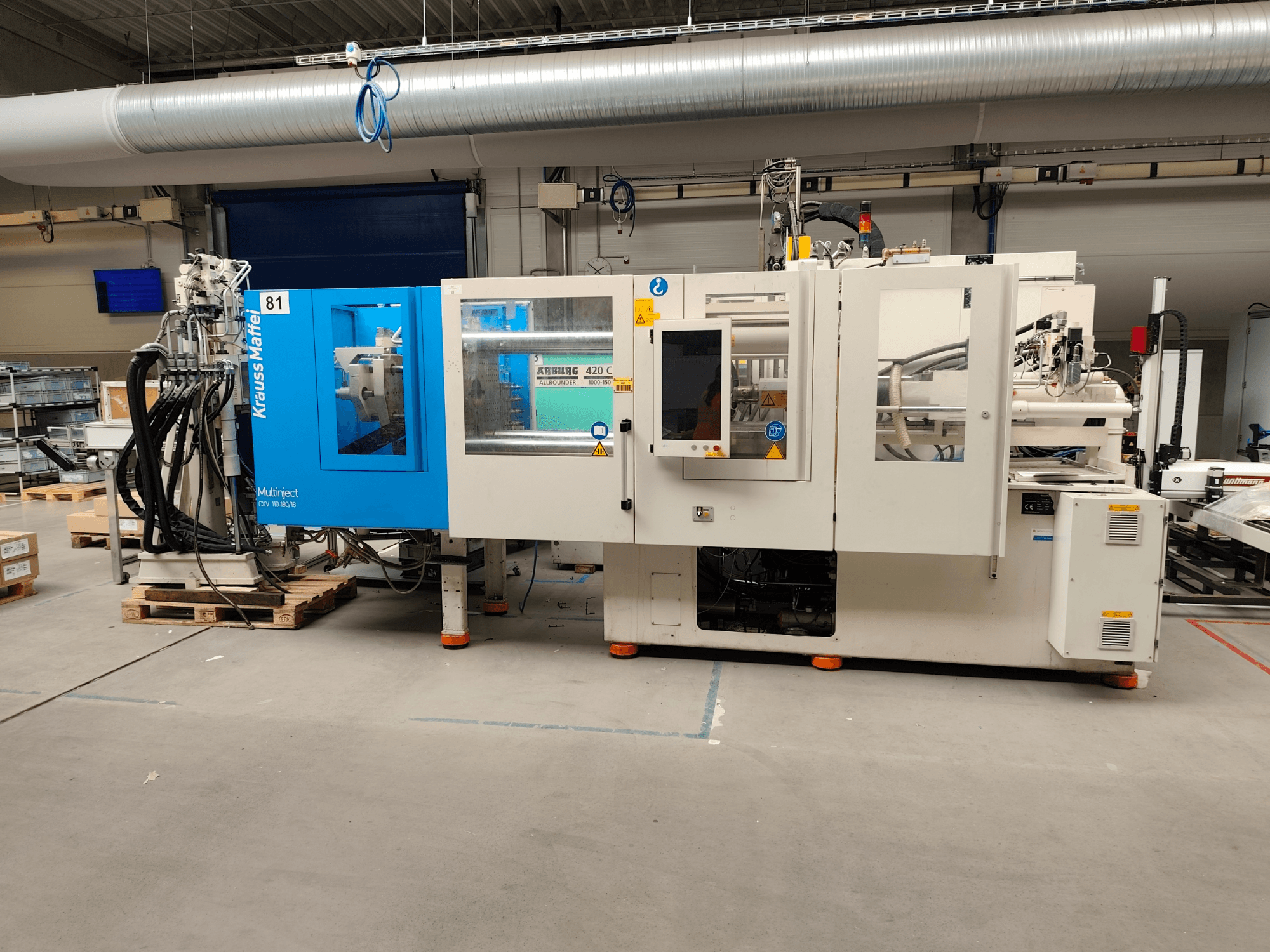 Hydraulische Spritzgießmaschine Krauss Maffei 110-180-180 CX V, Seitenansicht, mit blau-weißem Gehäuse, Bedienfeld und Anschlüssen.