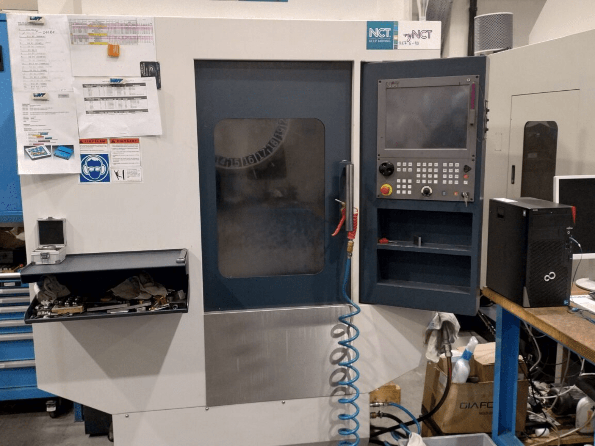 NCT CNC-Maschine, Vorderansicht, mit Bedienfeld mit numerischer Tastatur, Werkzeugen in der Schublade und Handbuch an der Wand.