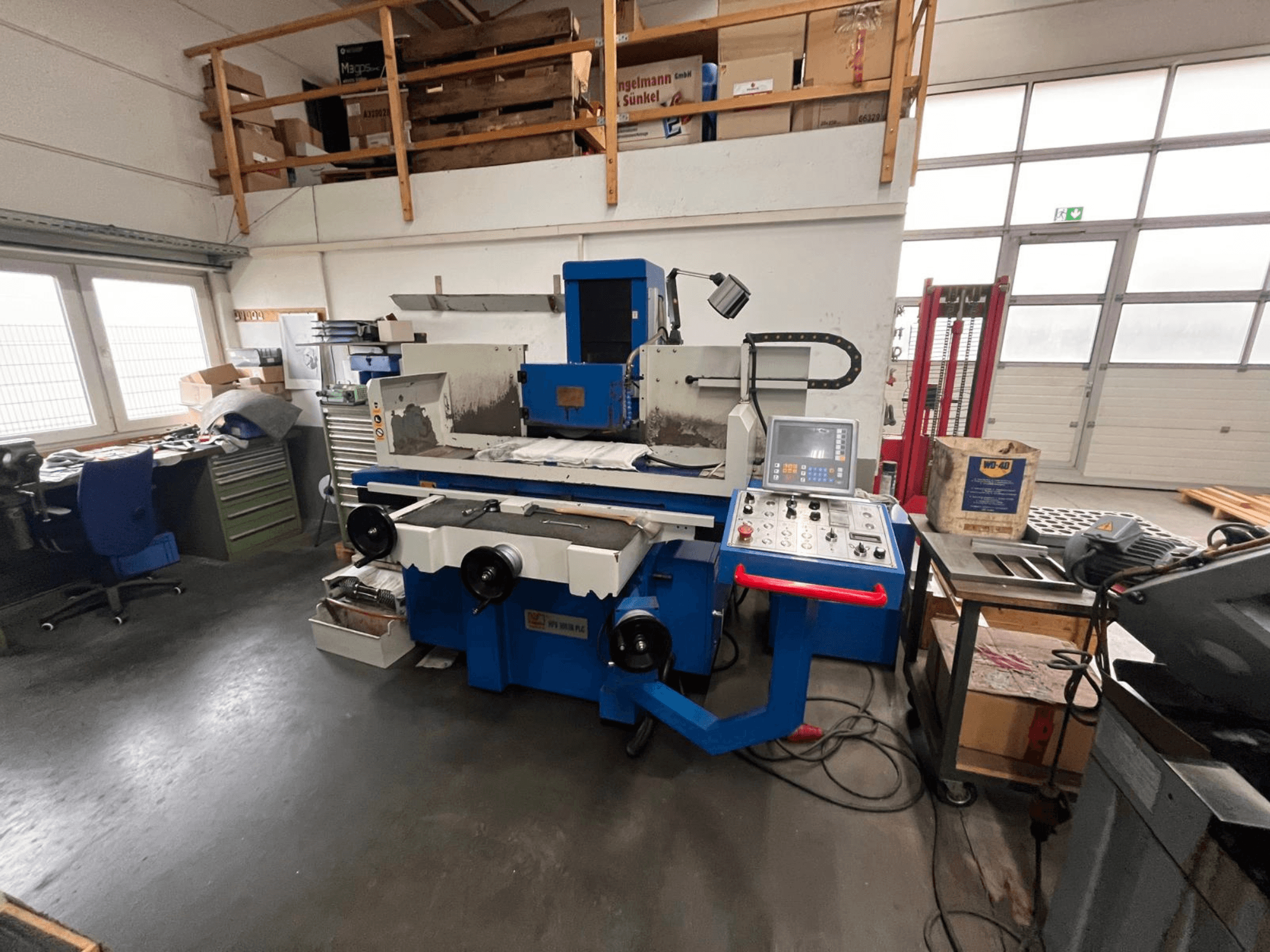 Blaue Flachschleifmaschine mit digitalem Bedienfeld, Vorderansicht, mit unübersichtlichem Arbeitsbereich und grünem Werkzeugschrank.