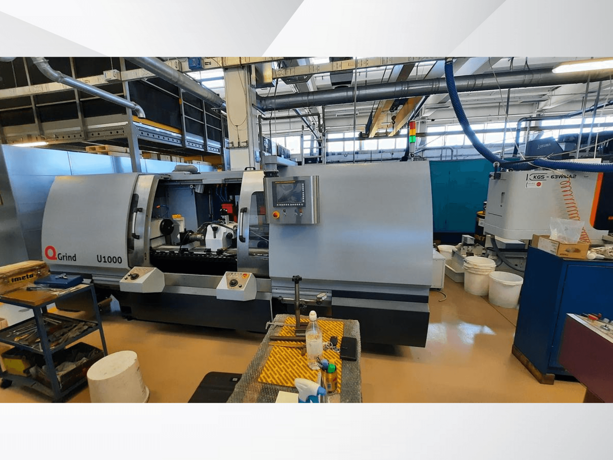 Q-Grind U1000 Maschine in einer Werkstatt, Frontansicht mit Bedienfeld und offenem Schneidbereich, umgeben von Geräten.
