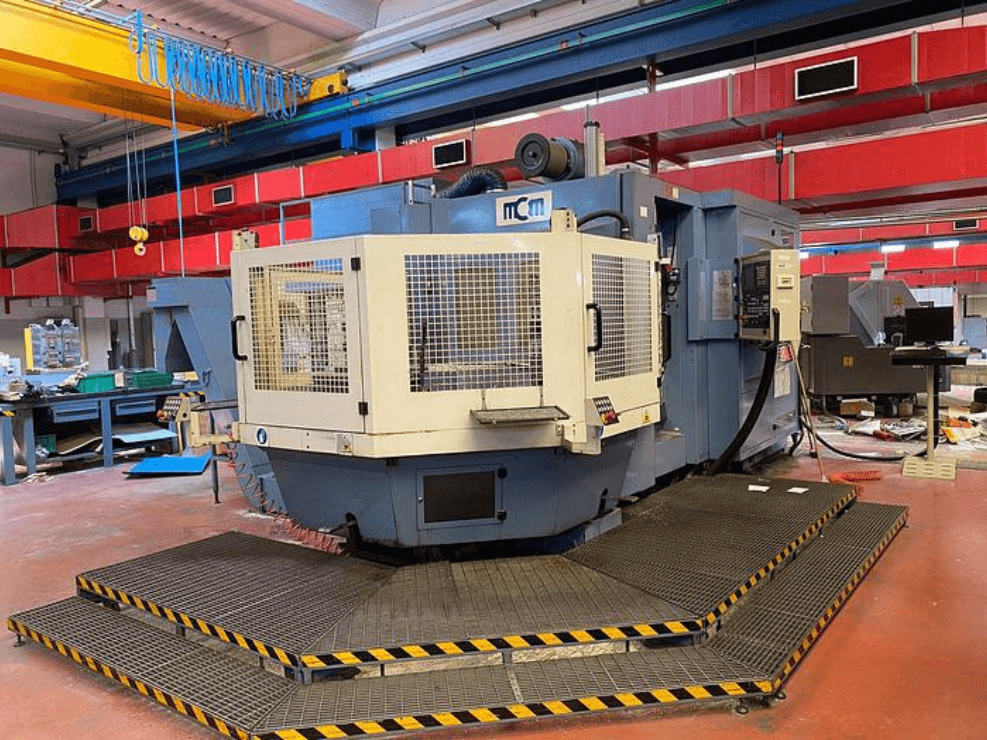 MCM-CNC-Maschine in einer Fabrik, Frontansicht mit Schutzkäfig, schwarze und gelbe Sicherheitsleisten, umgeben von Industrieanlagen.