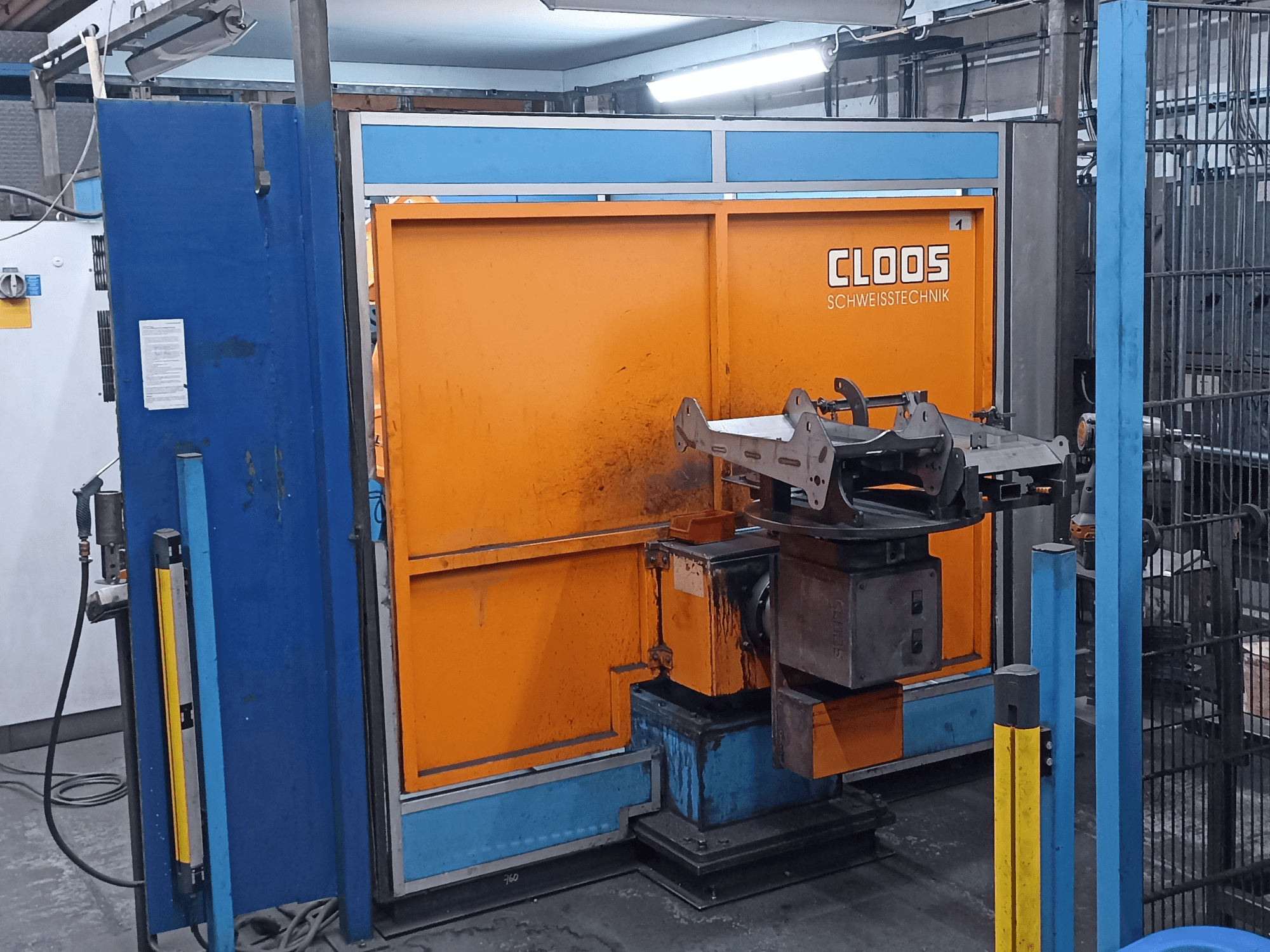 CLOOS Romat 320-Schweißroboter in einer Werkstatt, Vorderansicht, mit einem orangefarbenen Bedienfeld und einem Metallwerkstück auf einem drehbaren Sockel.