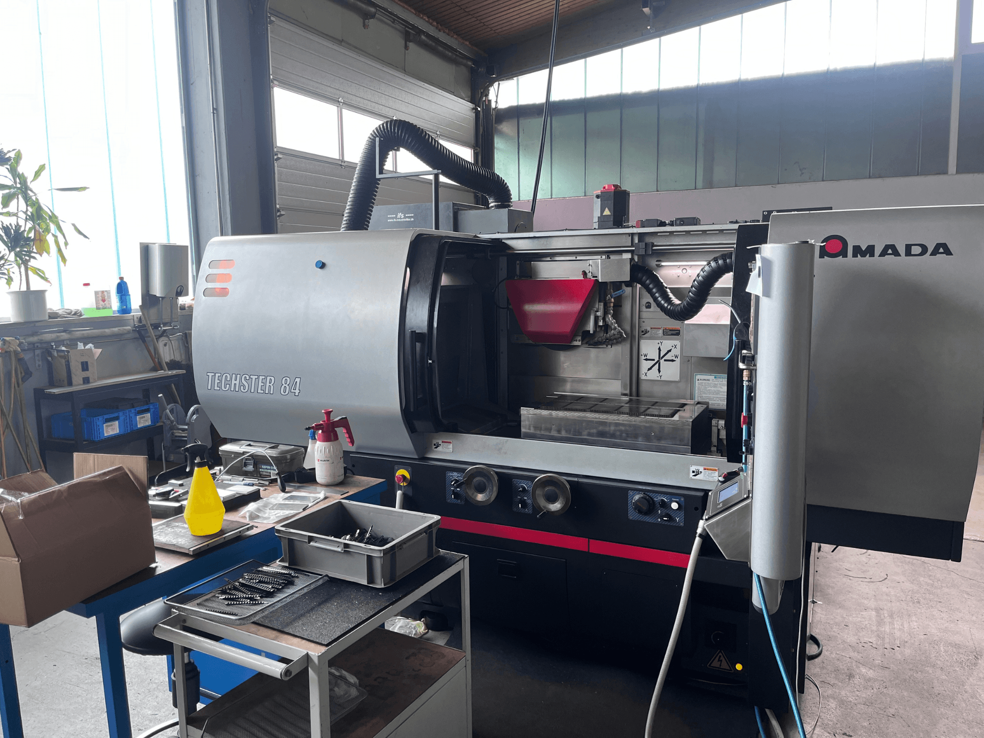 Eine AMADA Techster 84-Flachschleifmaschine in einer Werkstatt, gezeigt in einer Vorderansicht mit Bedienelementen und rotem Trichter.