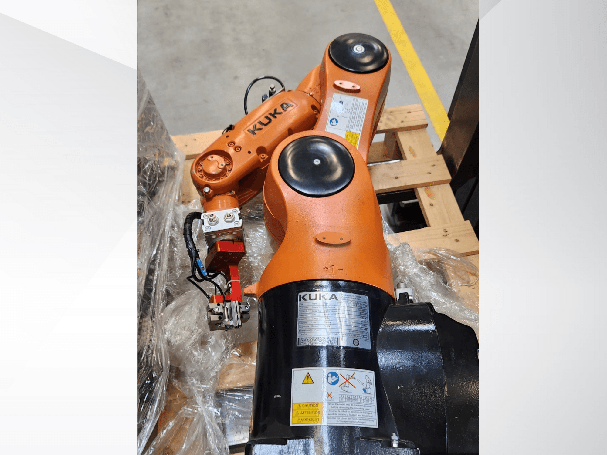 KUKA-Roboterarm in orangefarbener Ausführung, Seitenansicht, mit Greifer und Steuerleitungen auf einer Holzpalette.