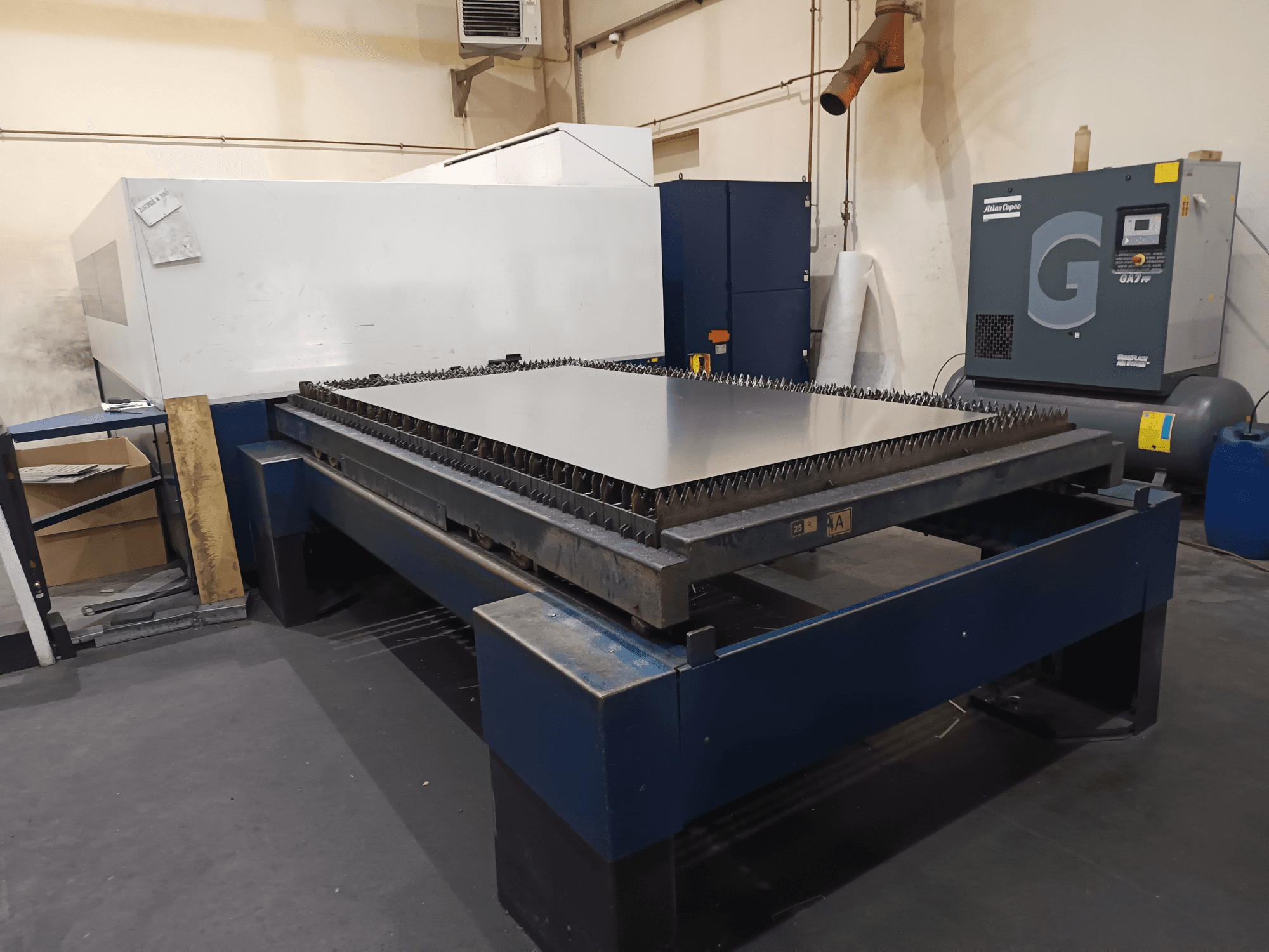 Industrielle Schneidemaschine mit Metalltisch, Vorderansicht. Mit scharfen Kanten, Luftkompressor auf der rechten Seite und Stahlrahmen.