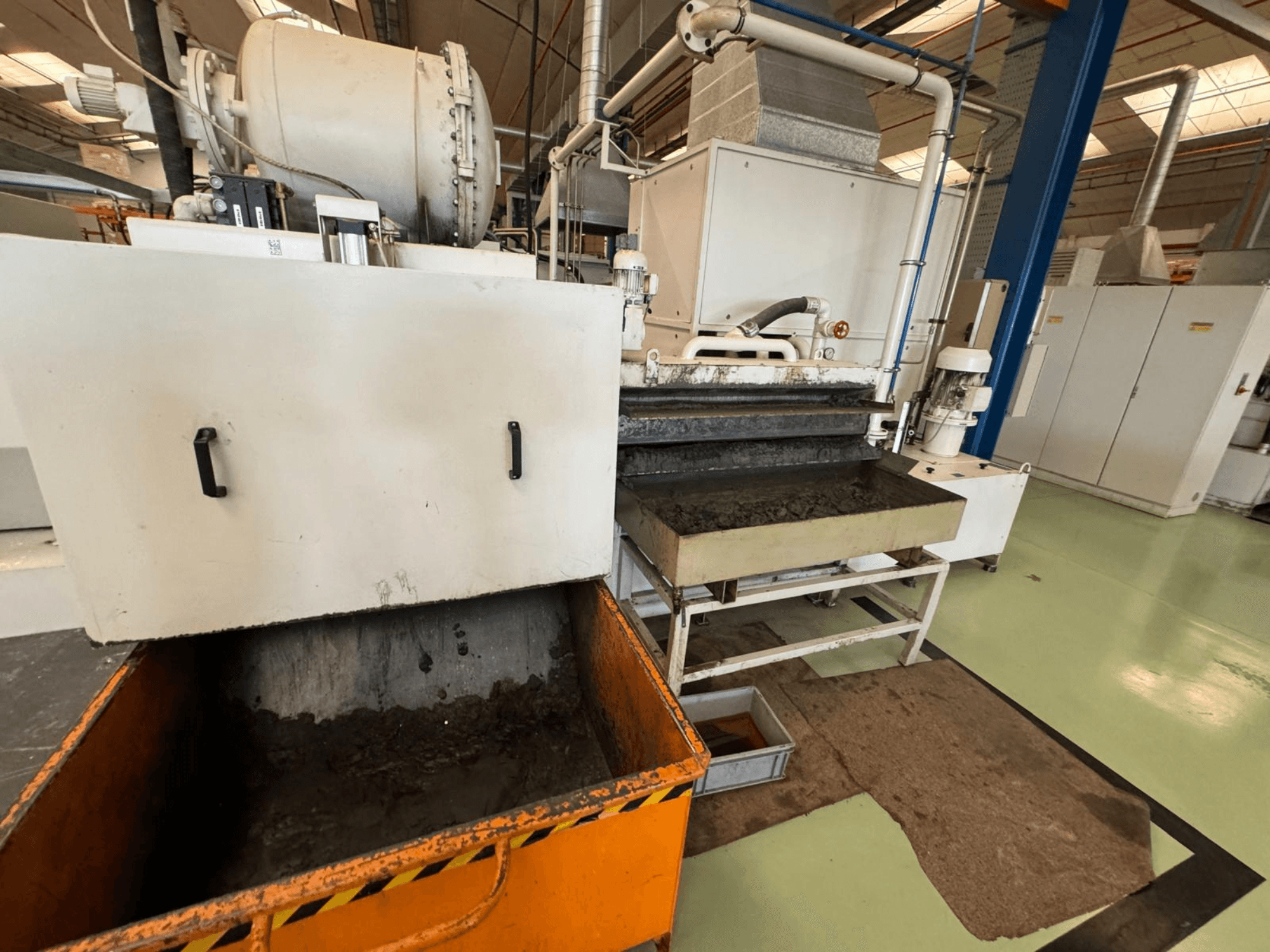 Industrielle Verarbeitungsanlage mit orangefarbenem Abfallbehälter, Vorderansicht; enthält einen Motor, Förderbänder und einen Wartungsbereich.