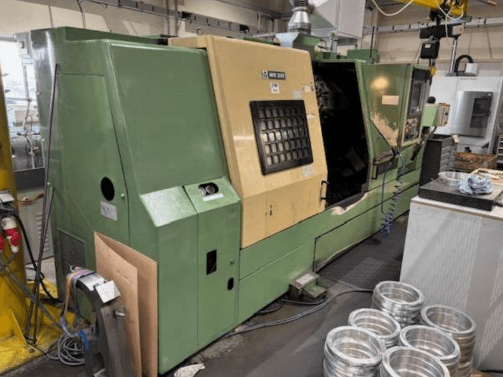 Grüne CNC-Drehmaschine von Mazak, von vorne gesehen, mit einem Bedienfeld und bearbeiteten Aluminiumteilen auf einem Arbeitstisch.