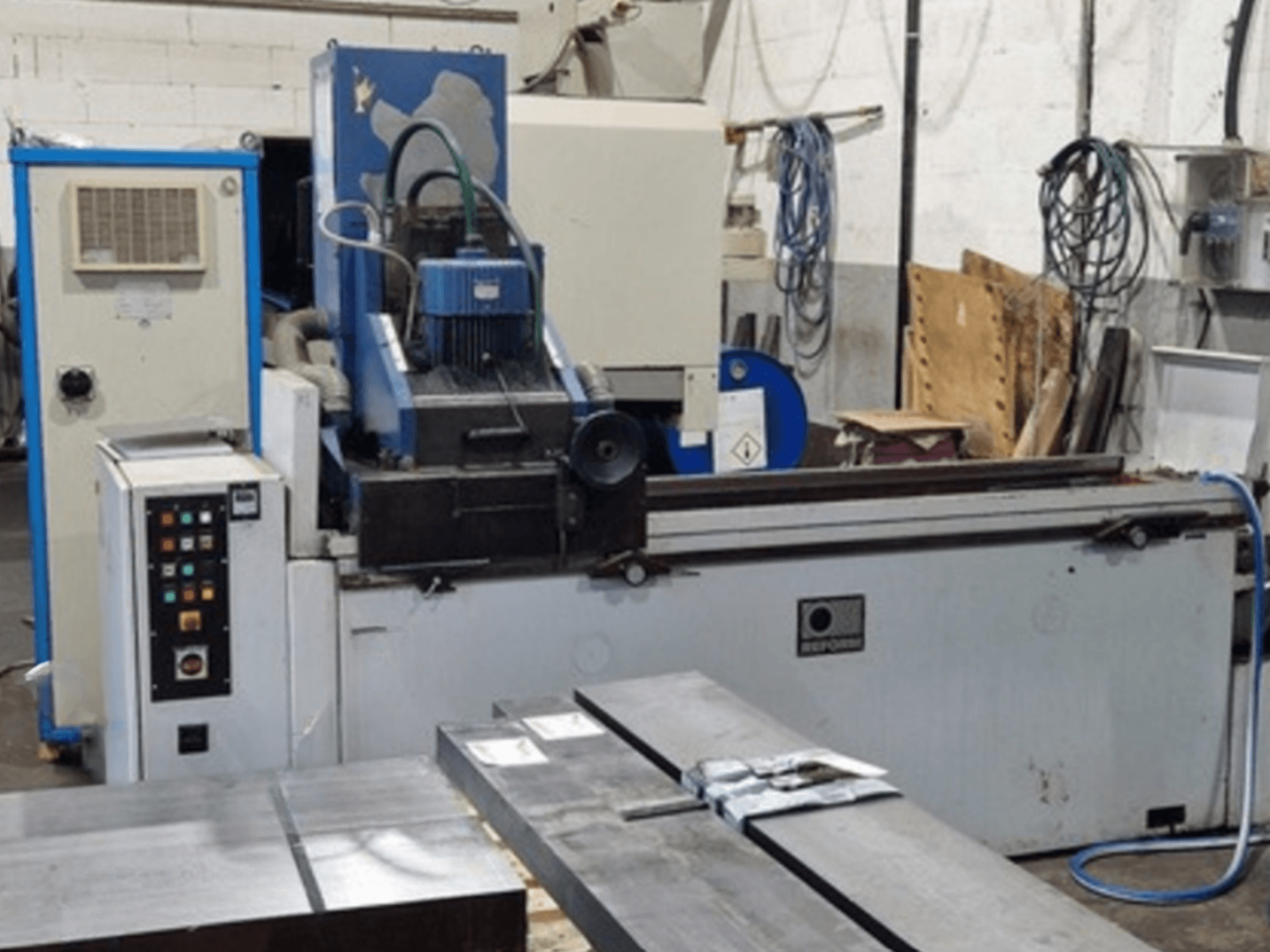 Reform-Flachschleifmaschine in Vorderansicht, mit Bedienfeld, Schleifbereich und Metallwerkstücken auf dem Tisch.