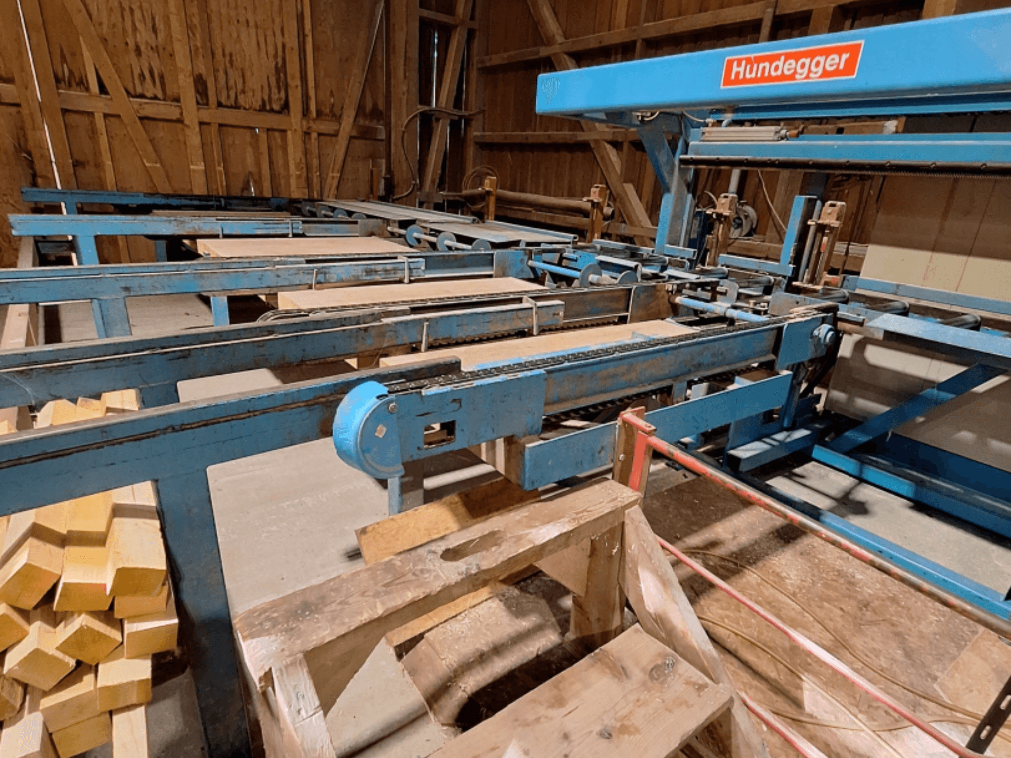 CNC-Holzbearbeitungszentrum HUNDEGGER K2-A in einer Scheune, mit blauem Metallrahmen, Fördersystem und gestapelten Holzbalken.