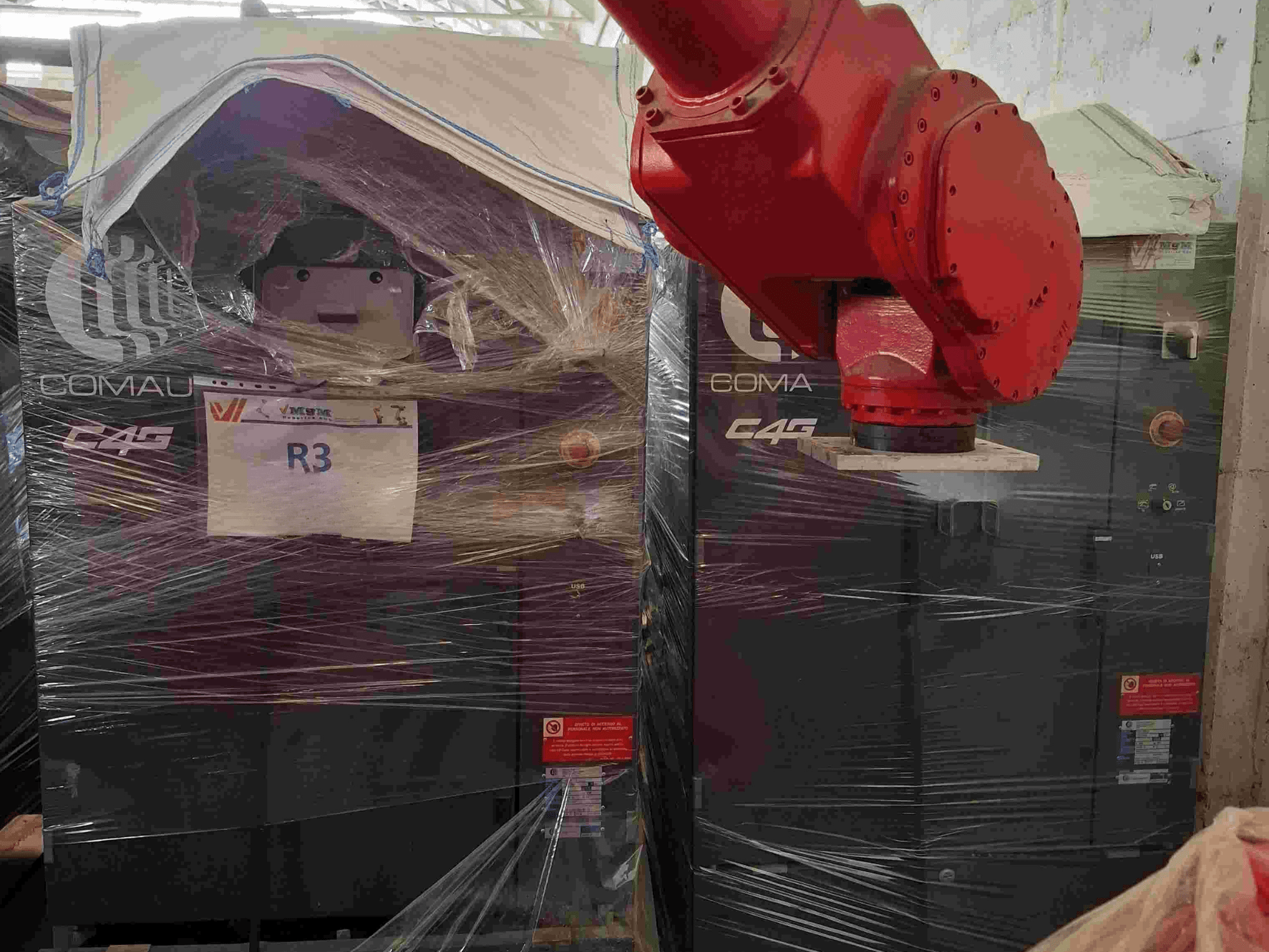 Comau C4G-Roboterarm von vorne gesehen, in Plastik verpackt, mit der Aufschrift "R3" und sichtbaren Warnaufklebern an der Maschine.