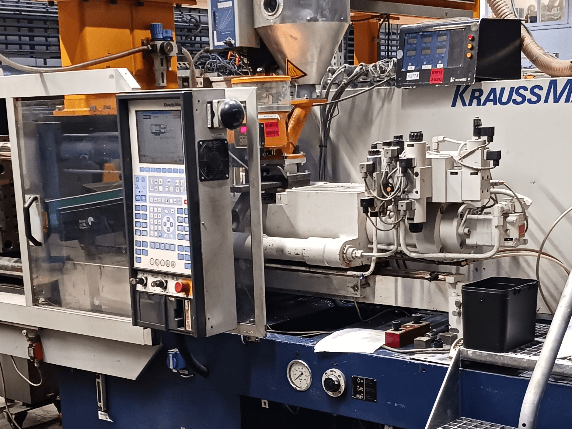 Spritzgießmaschine von KraussMaffei mit Bedienpult, Hydraulikaggregaten und Werkzeugen im Vordergrund, Vorderansicht.