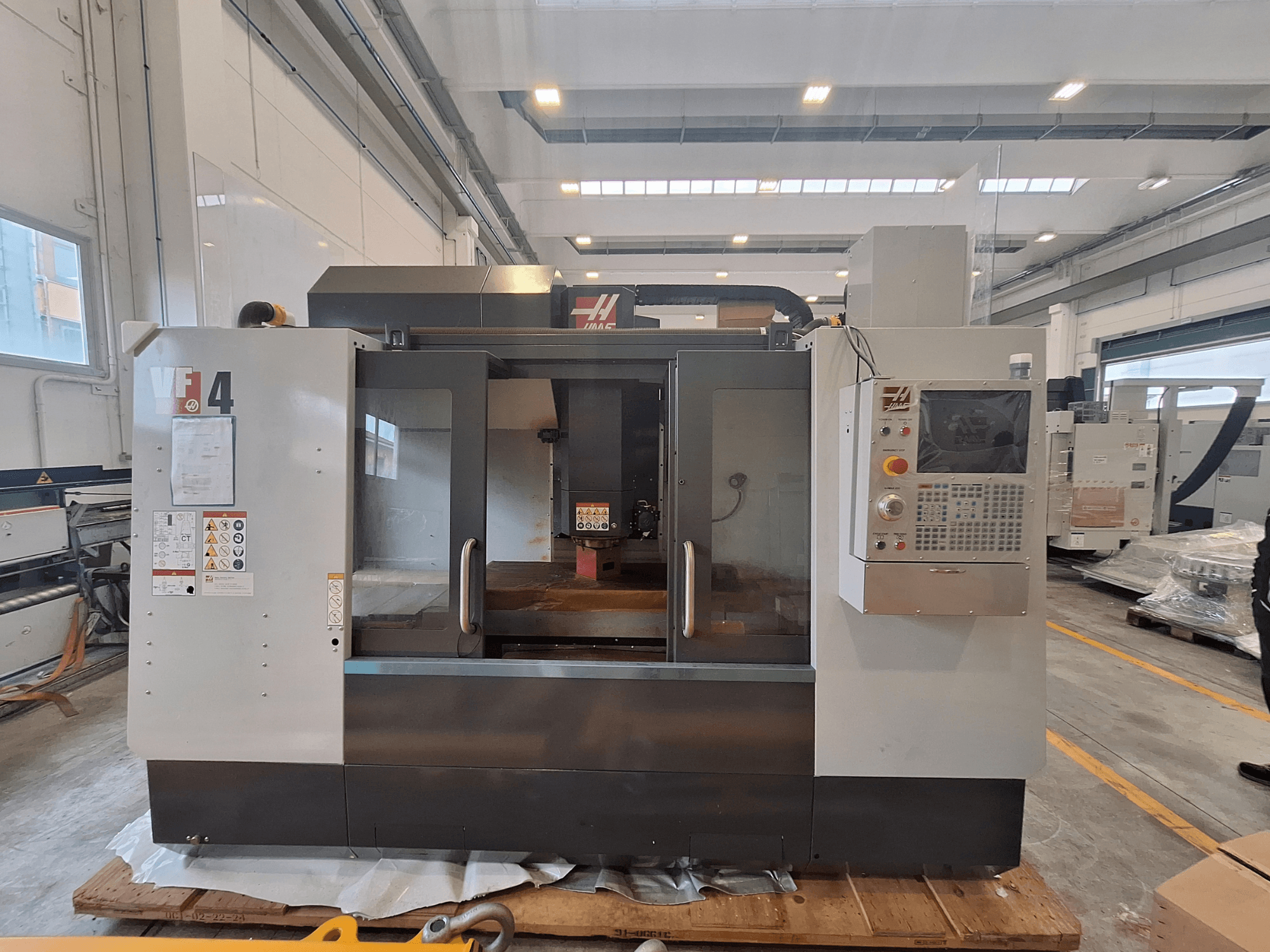 Vertikales Bearbeitungszentrum HAAS VF-4, Vorderansicht, mit Blick auf das Bedienfeld, den Arbeitsbereich und die Sicherheitshinweise in einer Lagerumgebung.