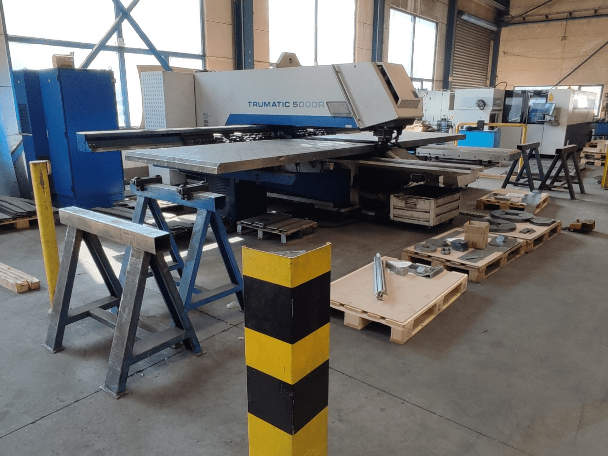 CNC-Stanzmaschine TRUMPF Trumatic 5000 R in einer Werkstatt, Seitenansicht mit Werkzeugen und Materialien auf Paletten in der Nähe.