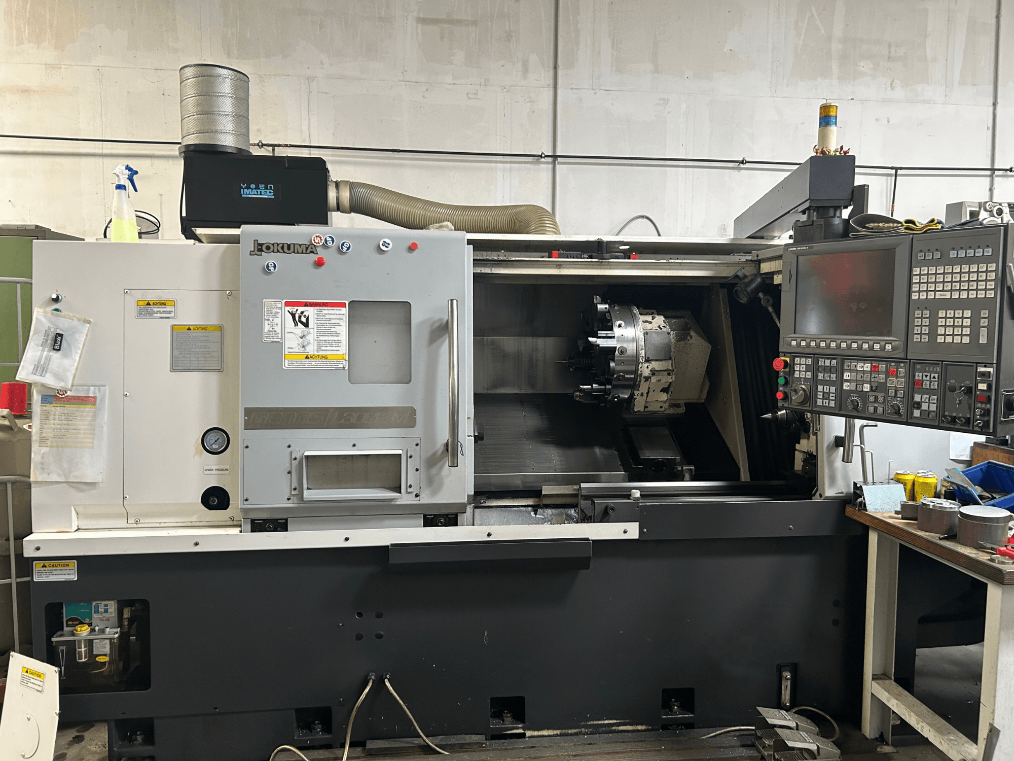 Frontansicht einer Okuma Genos L300E-M Horizontaldrehmaschine mit Bedienfeld, sichtbarer Spindel und verschiedenen Einrichtwerkzeugen.