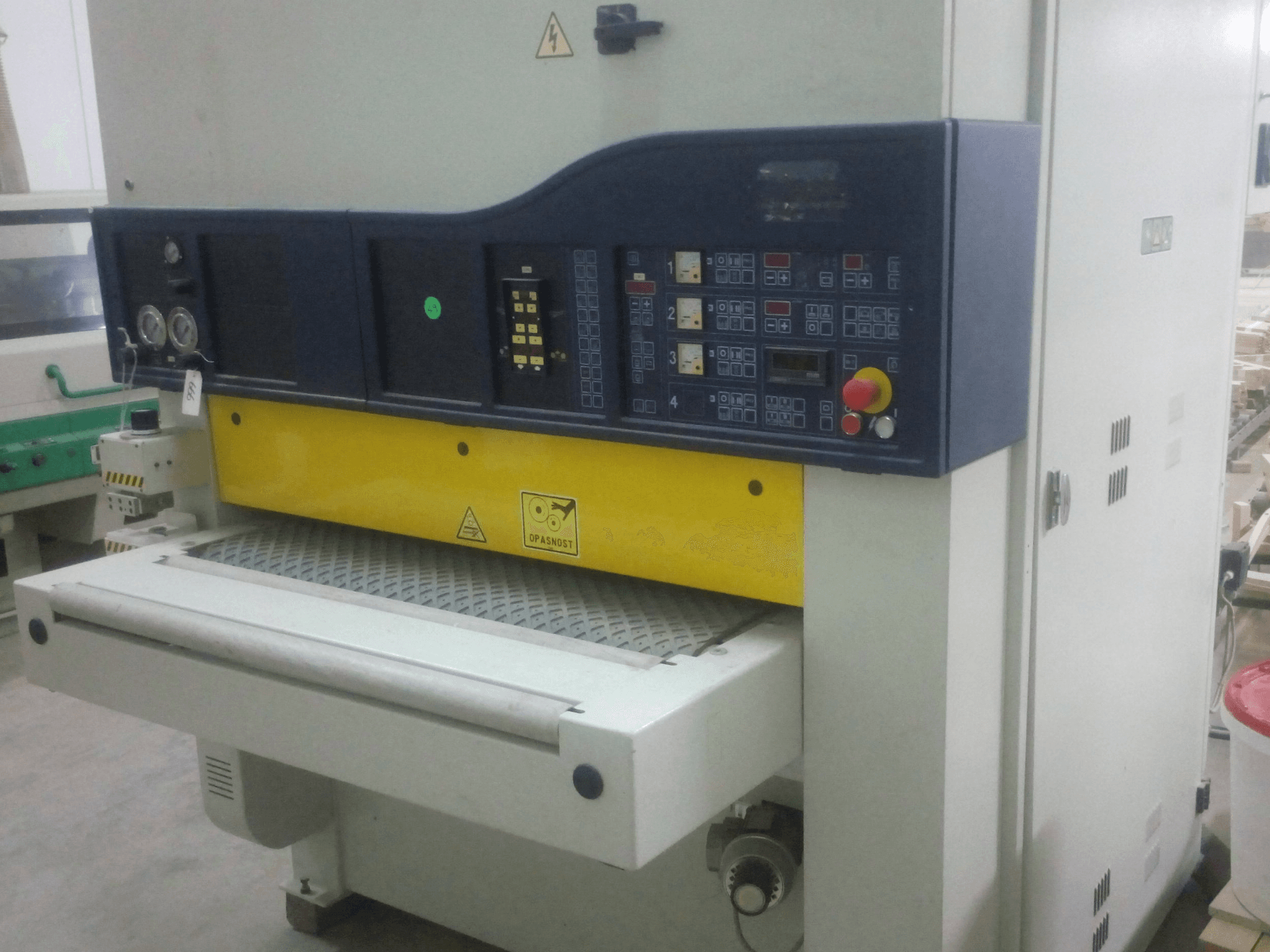 Industrielle Schleifmaschine mit Schalttafel, gelbes und blaues Gehäuse, Vorderansicht, mit Messgeräten, Schaltern und Förderfläche.