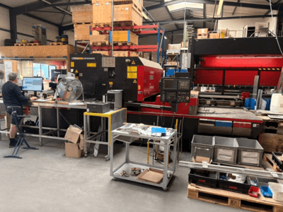 Eine CO₂-Laserschneidmaschine vom Typ AMADA LC-2415αIII in einer Werkstattumgebung, im Vordergrund ist ein Computerarbeitsplatz zu sehen.