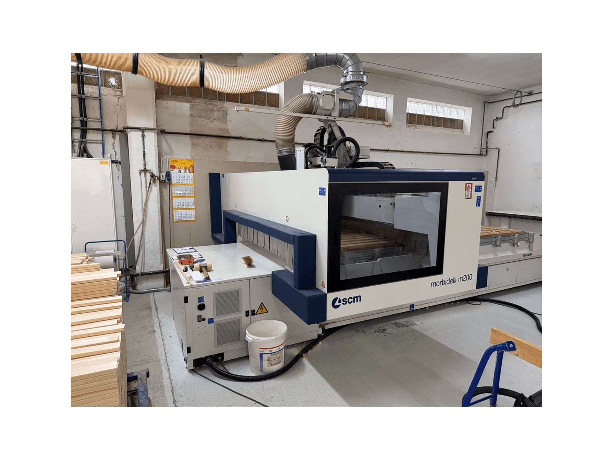 CNC-Bearbeitungszentrum SCM Morbidelli M200F in einer Werkstatt, Seitenansicht mit Holzstücken auf dem Tisch und einer Lüftungsanlage darüber.