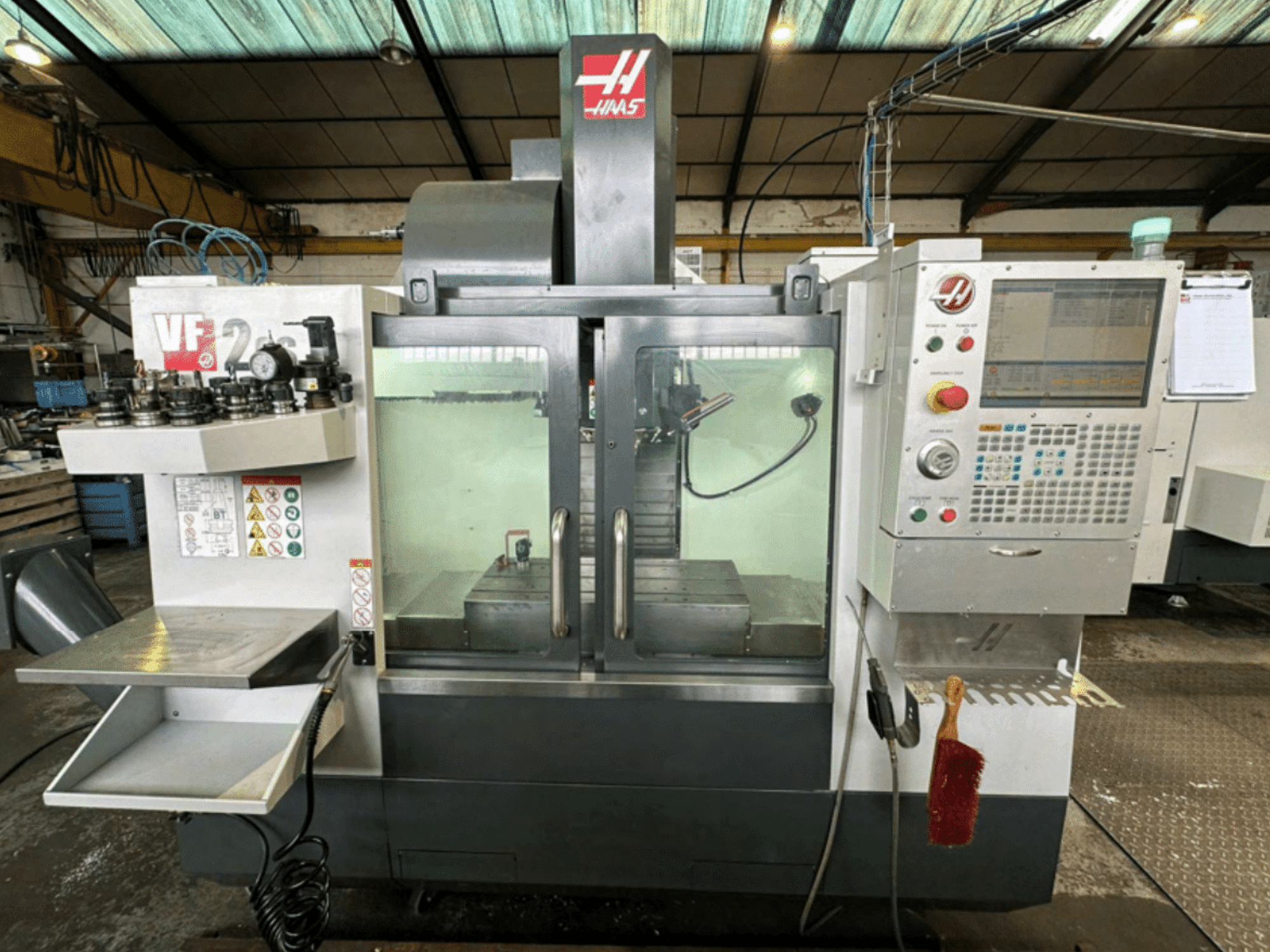 Vorderansicht der Haas VF2 CNC-Maschine mit Bedienfeld, Werkzeughaltern und Arbeitsbereich mit Sicherheitsetiketten und einer Bürste.